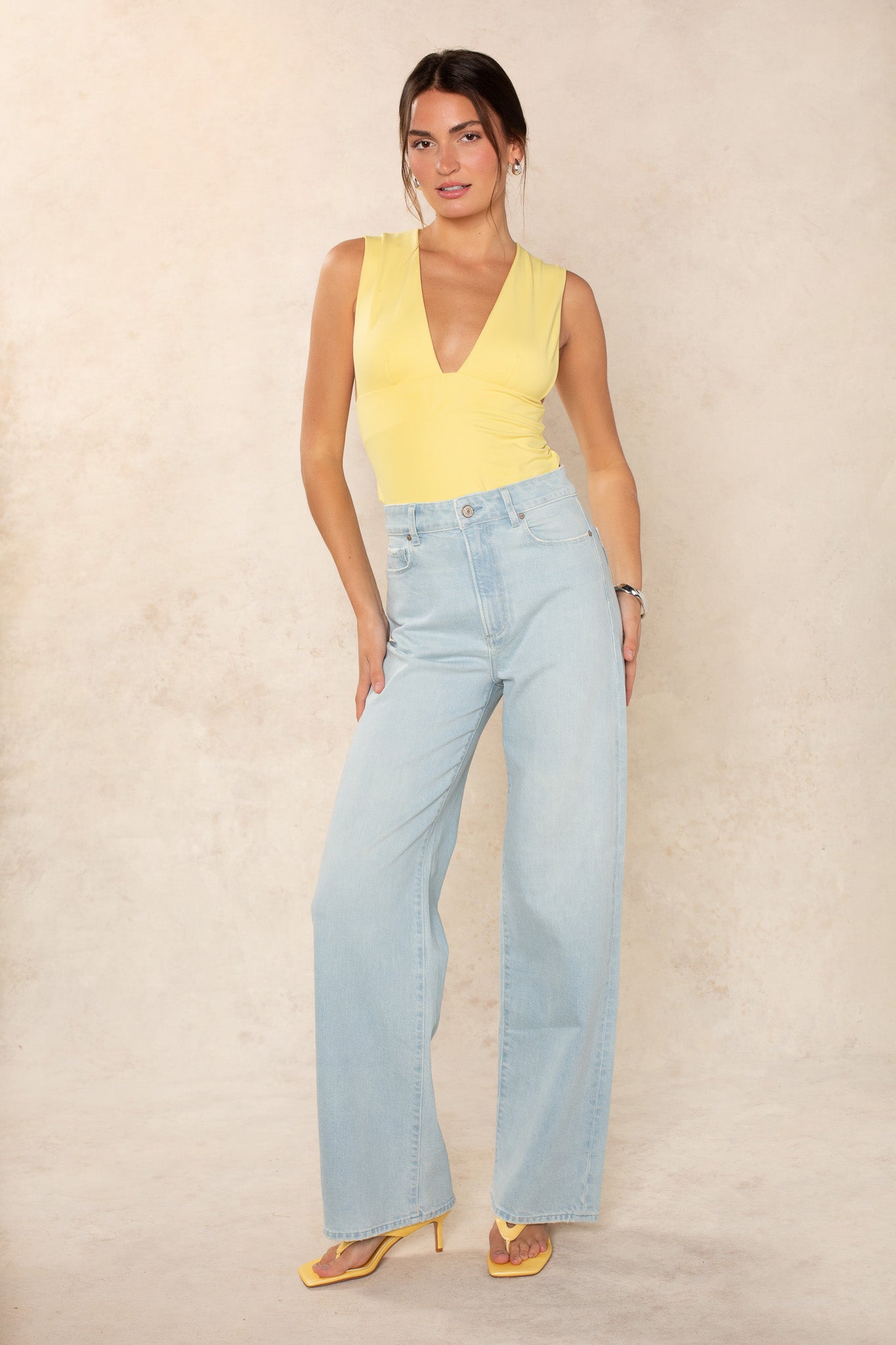 A woman with tied-back dark hair and minimal accessories stands against a neutral background wearing Georgie x Paige’s Anessa 31 Clean Hem - Fleur De Mer pale blue high-rise wide leg jeans, a yellow sleeveless V-neck top, and yellow heeled sandals.