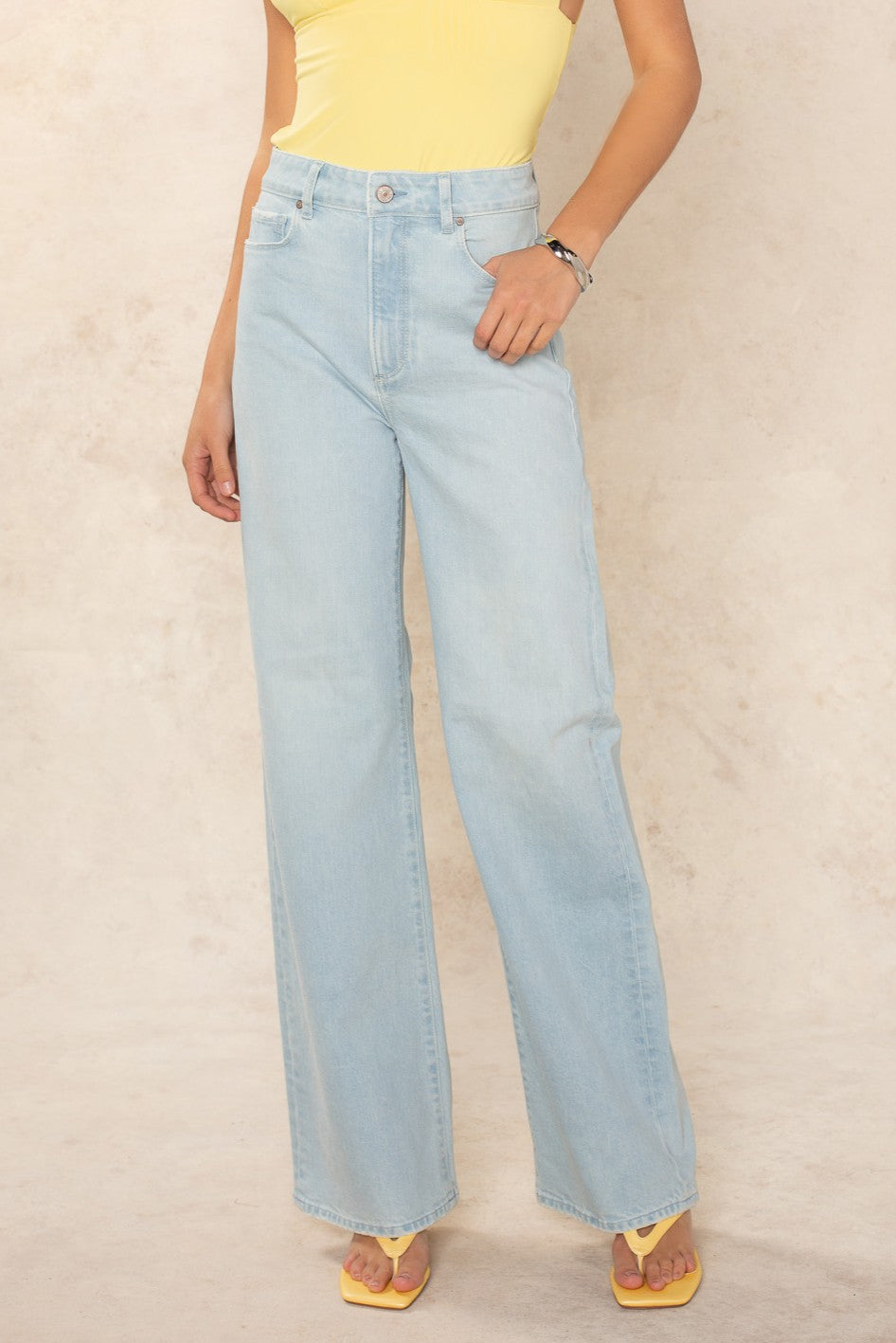 Wearing the Georgie x Paige Anessa 31 Clean Hem - Fleur De Mer pale blue high-rise wide leg jeans, a sleeveless yellow top, yellow sandals, and a bracelet, this person stands against a light beige background.