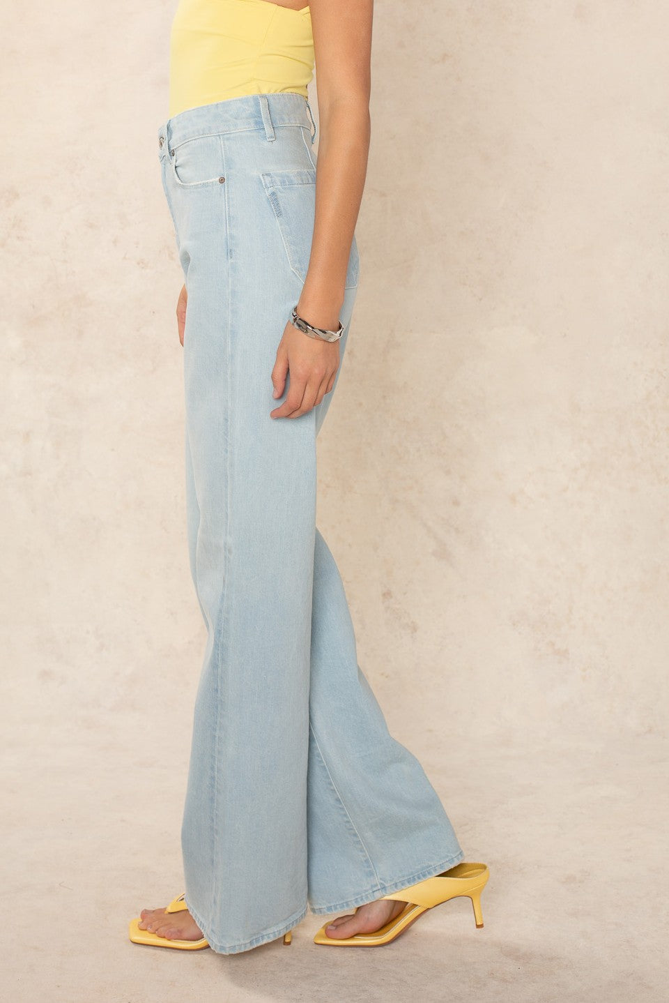 A person stands sideways against a beige background wearing Georgie x Paige’s Anessa 31 Clean Hem - Fleur De Mer pale blue denim high-rise wide leg jeans, a yellow sleeveless top, yellow heeled sandals, and a silver bracelet.