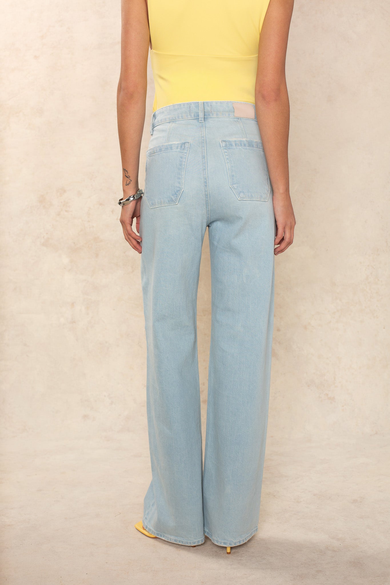 The lower half of a person is shown wearing Georgie x Paige's Anessa 31 Clean Hem - Fleur De Mer pale blue high-rise wide leg jeans, paired with a sleeveless yellow top against a neutral textured beige background.