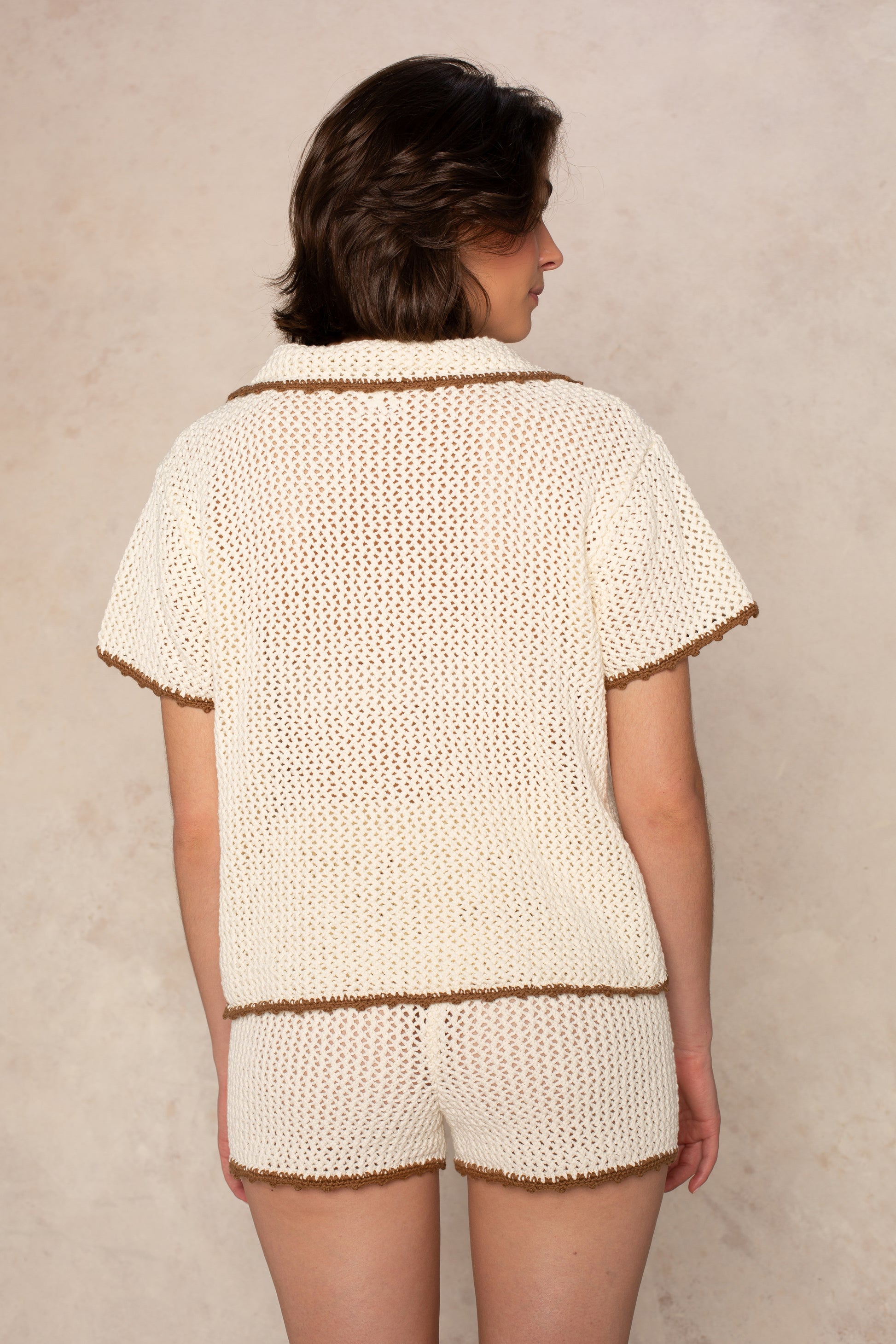 A person with short brown hair stands with their back to the camera, wearing the Isla Crochet Contrast Piping Coverup Top and matching shorts, both featuring brown trim, against a light textured background.