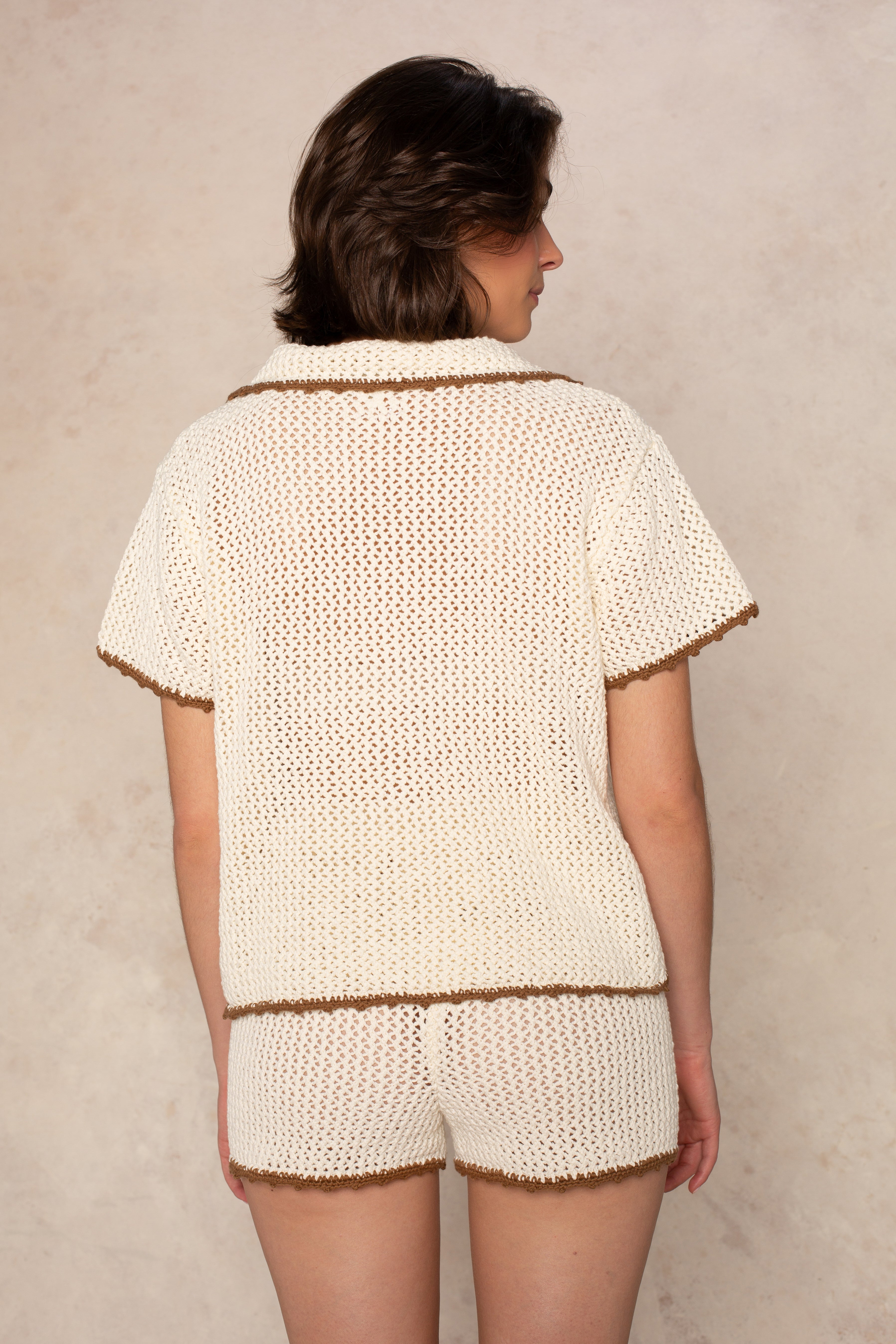 Against a light beige textured background, a person with short brown hair wears Isla’s Crochet Contrast Piping Coverup Short—a lightweight cream crochet set with brown piping detail—seen from behind.