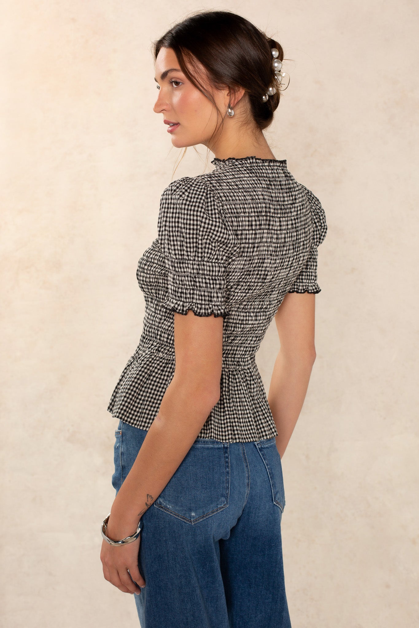 A woman in profile wears the Georgie Lynn Gingham Short Sleeve Top with puffed sleeves and blue high-waisted jeans. Her brown hair is styled up with loose strands, set against a neutral background.