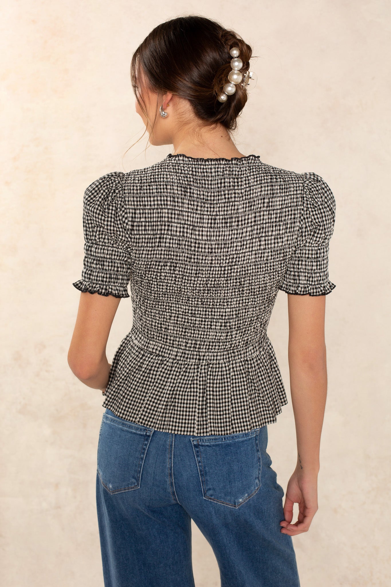 A woman with brown hair in a low bun styled with pearl pins wears the Georgie Lynn Gingham Short Sleeve Top and blue high-waisted jeans, standing with her back to the camera against a beige background.