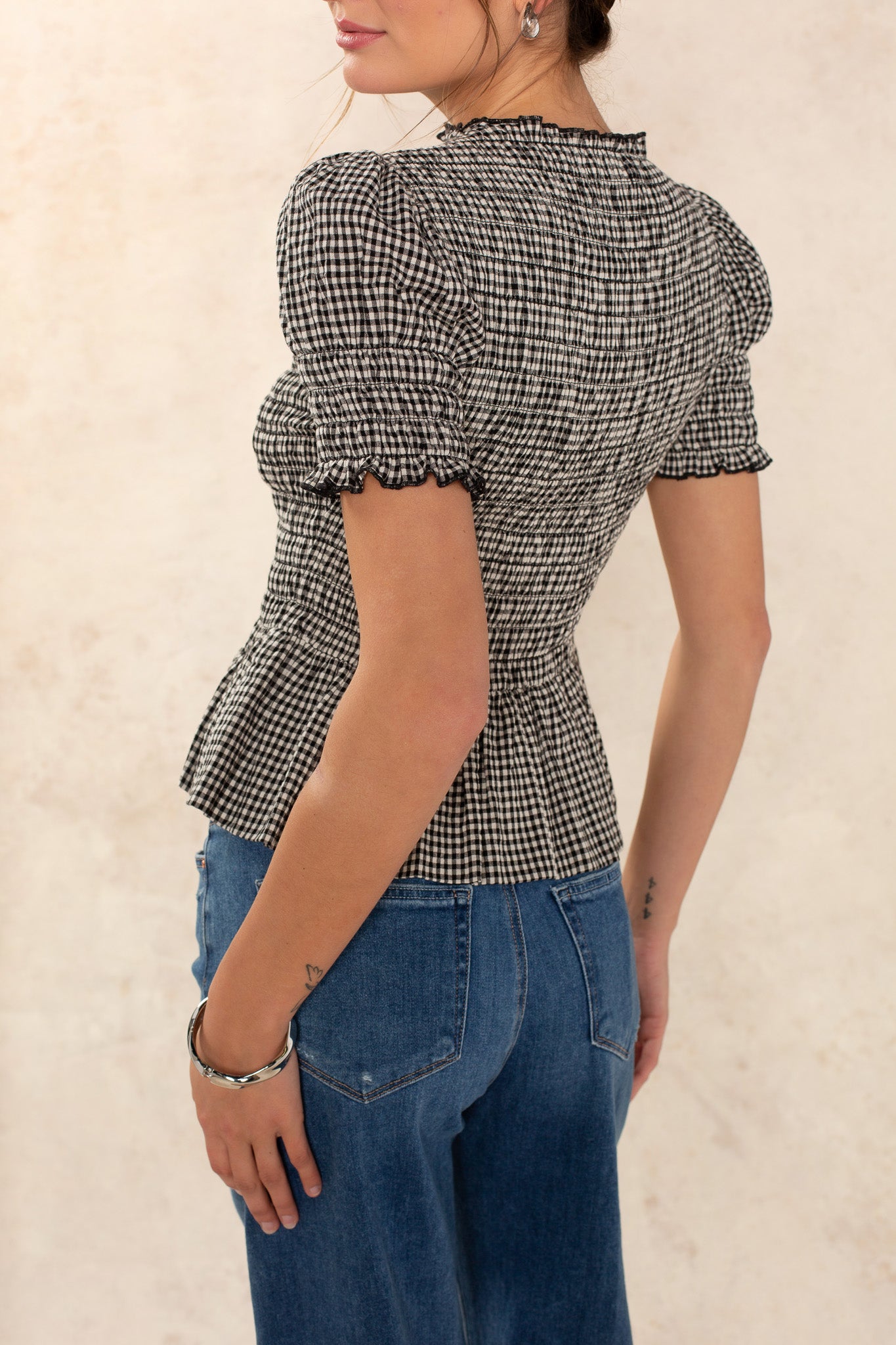 A woman models the Georgie Lynn Gingham Short Sleeve Top, featuring black and white checks and puffed sleeves, paired with blue jeans. She stands sideways with her head slightly turned and her hair tied back.
