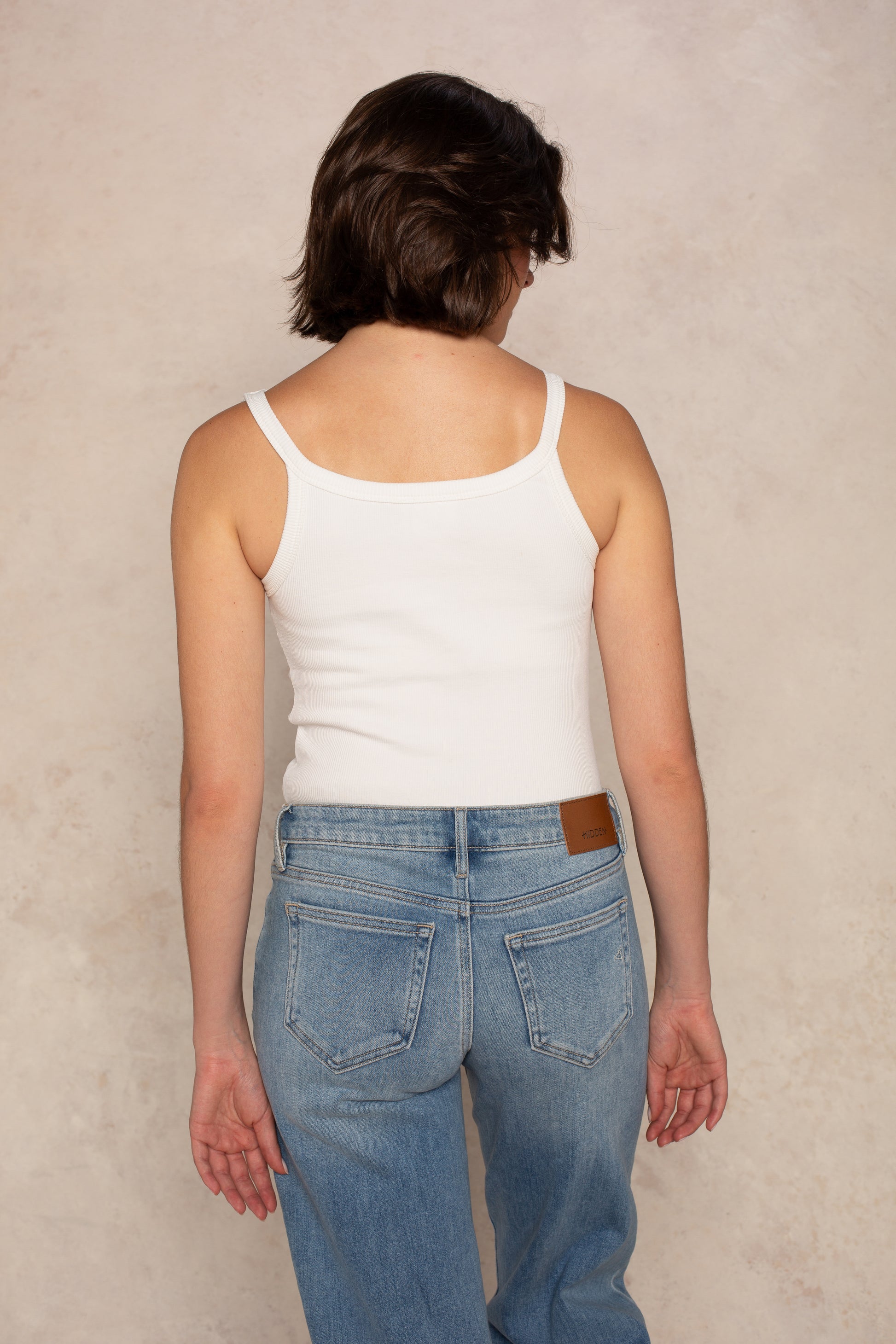 A person with short brown hair models the Isla Fish Embroidered Vintage Fit Tank, paired with blue jeans, standing with their back to the camera against a light neutral background.