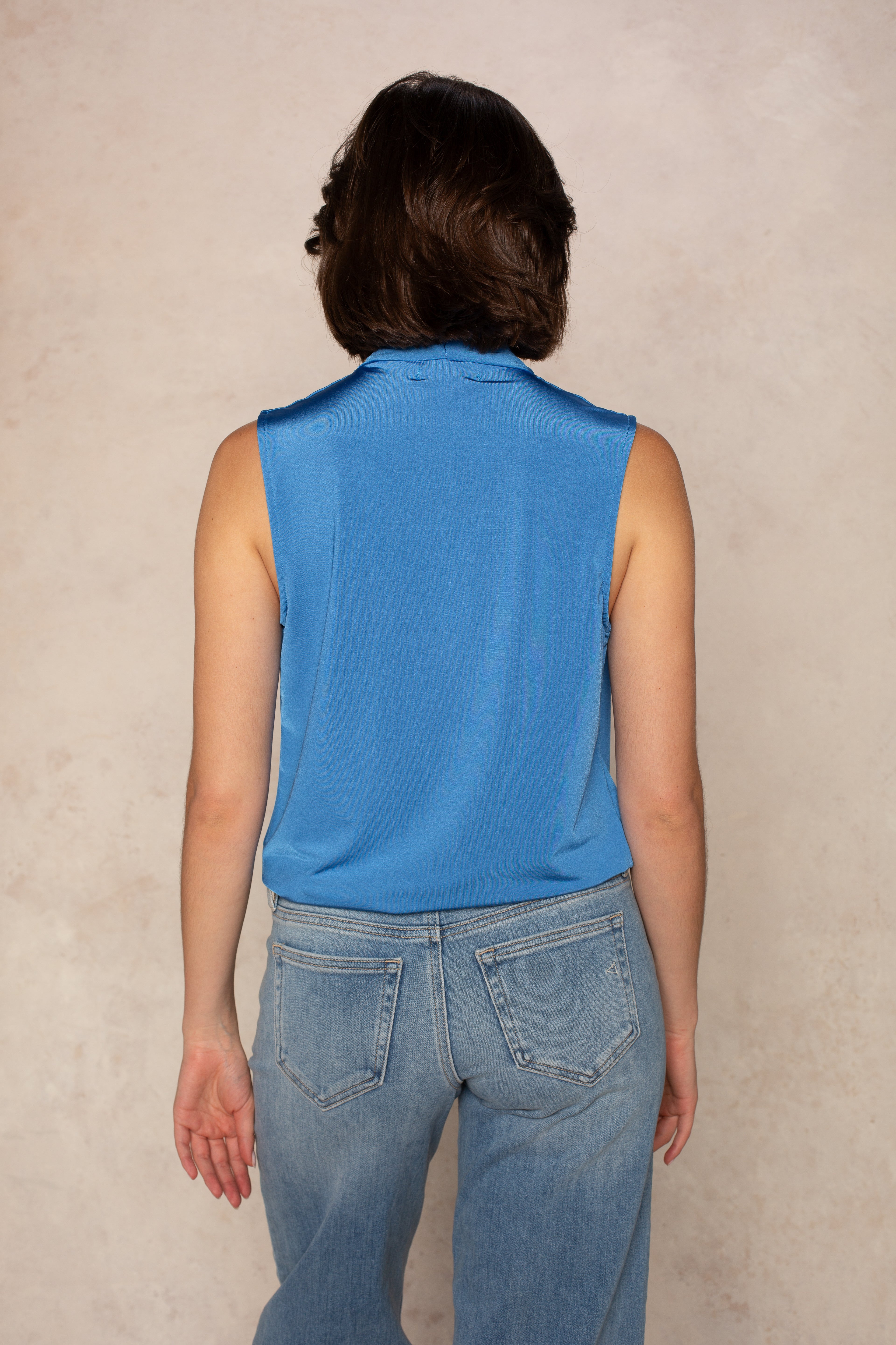 A person with short brown hair, seen from behind, wears the Georgie Gwen Sleeveless Wrap Top in blue with light blue jeans, standing against a neutral beige background.