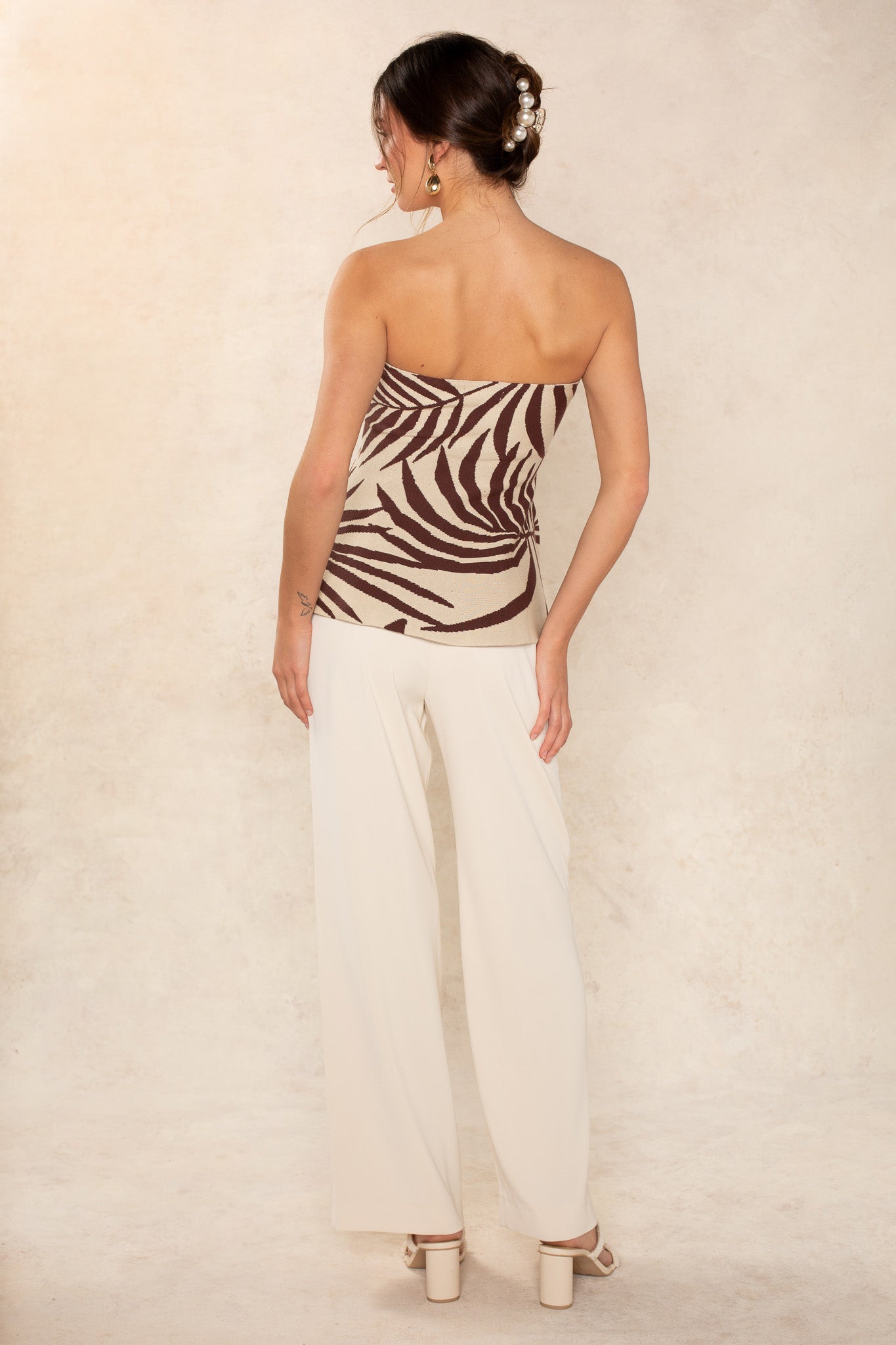 A woman stands facing away, wearing the Georgie Phoebe Peplum Tube with white wide-leg pants and heeled sandals. Her hair is styled in an updo with decorative hairpins, set against a neutral textured background.