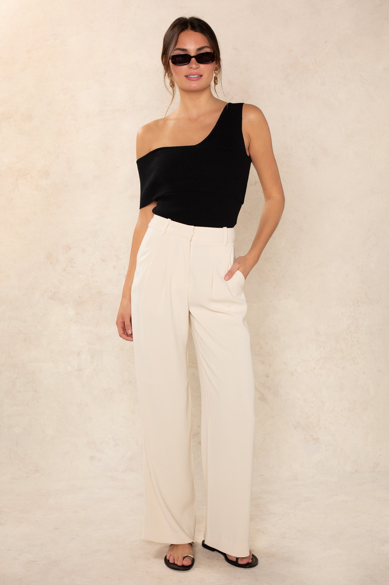 A woman wears the Georgie Lucy Off The Shoulder Top in black with cream high-waisted wide-leg trousers, black sandals, and sunglasses, posing against a light beige textured background.