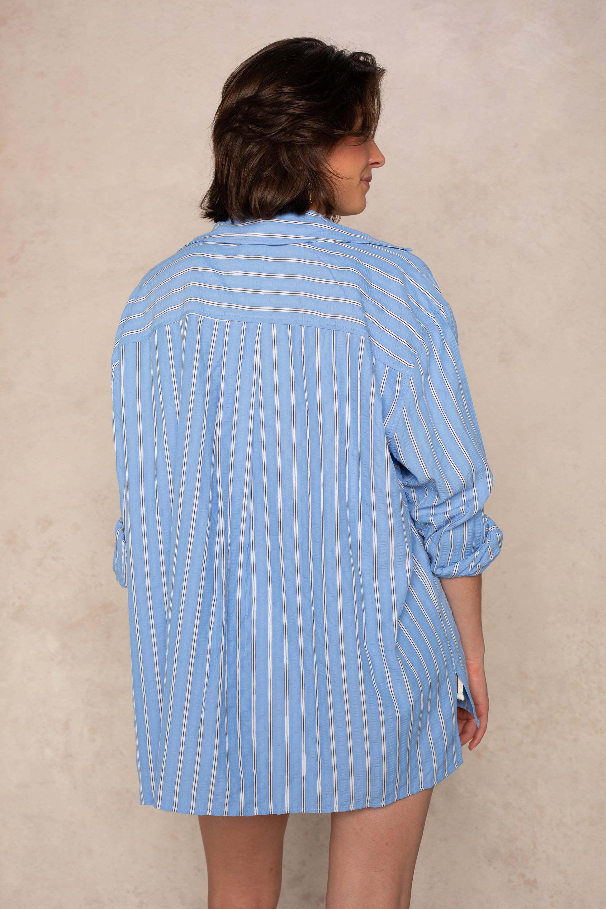 A person stands with their back to the camera, wearing the Isla Claire Striped Button Down Coverup Top with rolled sleeves and collared button-front, set against a neutral, textured background.