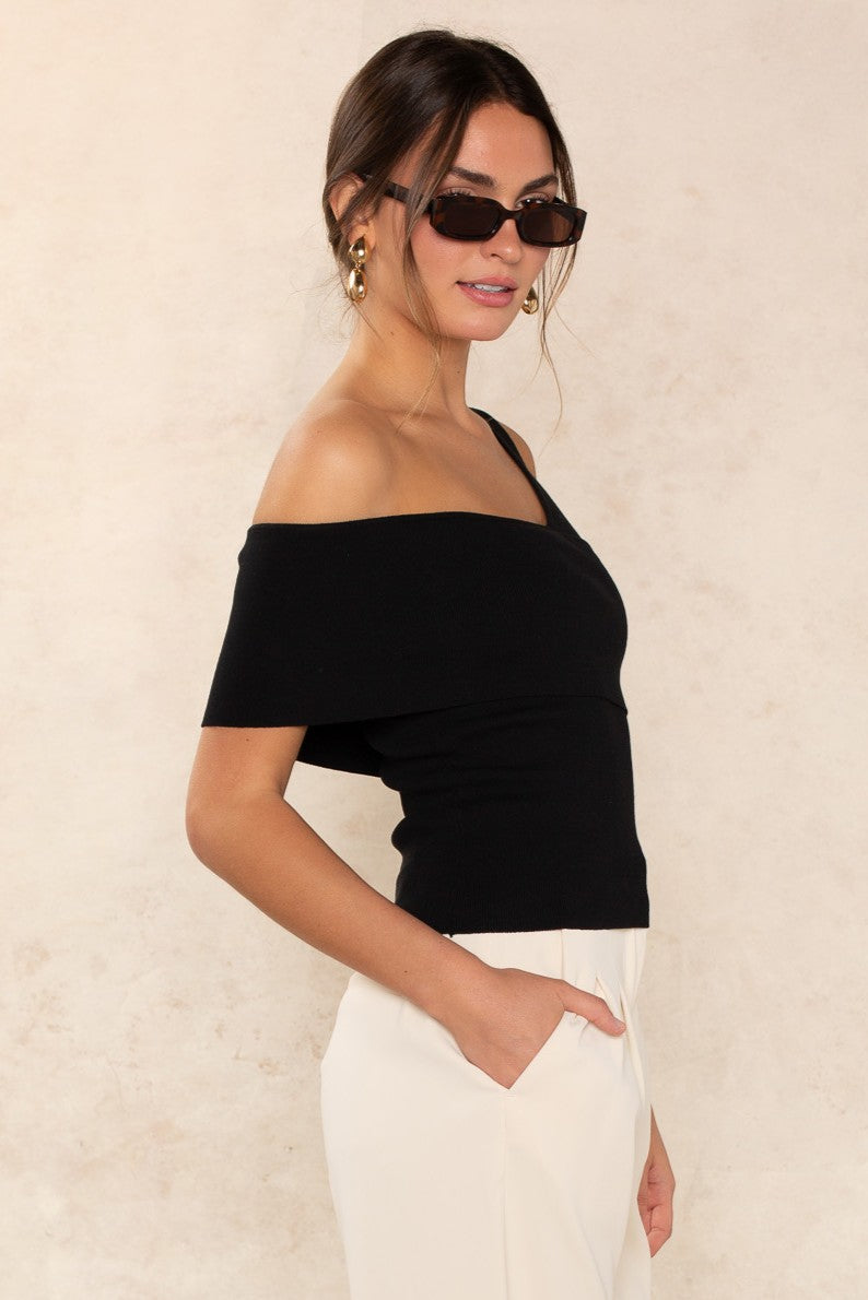A woman stands sideways against a light beige background, wearing the Georgie Lucy Off The Shoulder Top in black, paired with relaxed-fit white high-waisted pants, gold hoop earrings, and rectangular sunglasses, with one hand in her pocket.
