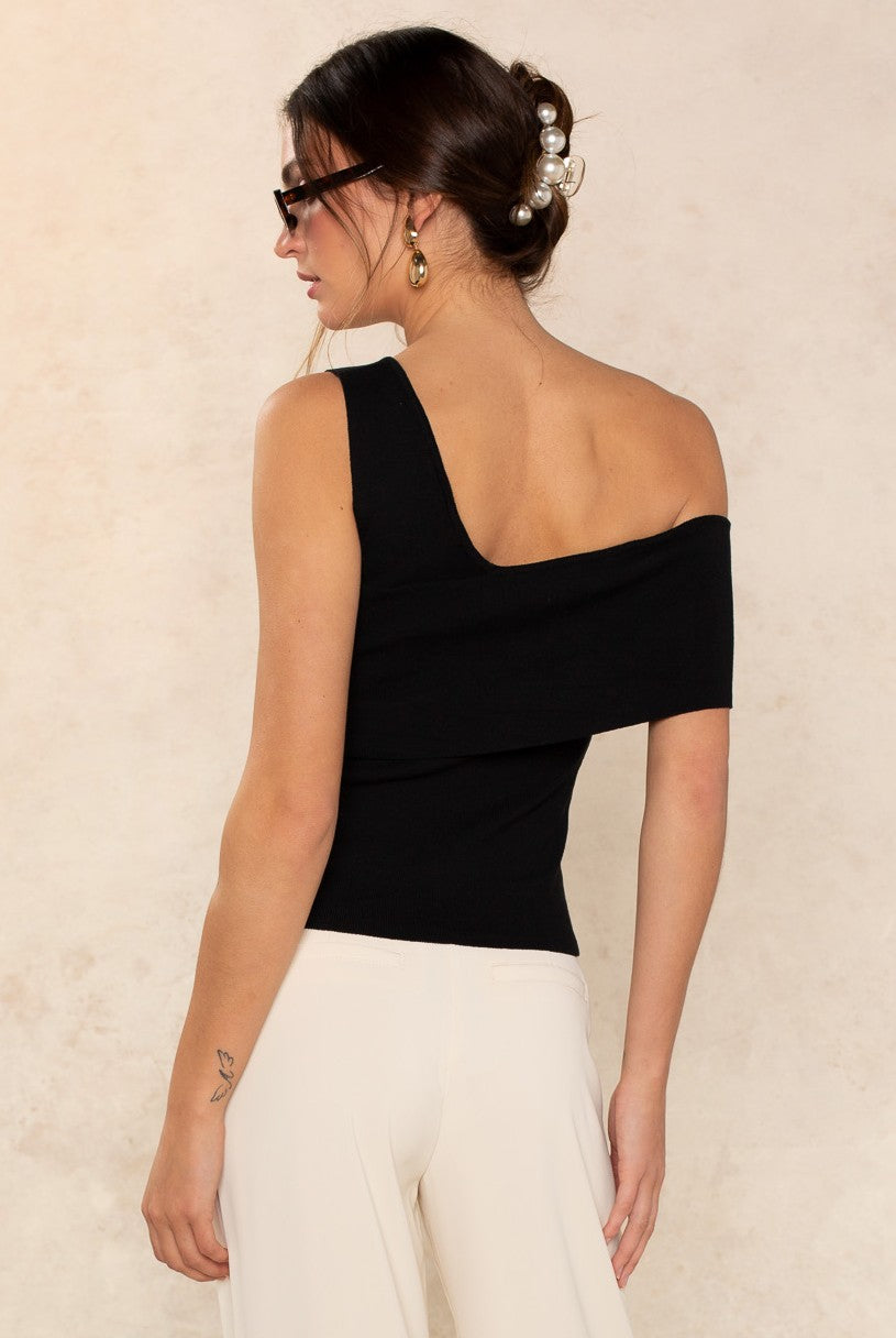 A woman with brown hair in a low pearl-adorned bun wears sunglasses, gold earrings, and the Georgie Lucy Off The Shoulder Top with cream wide-leg pants, standing against a light beige background and shown from the back.