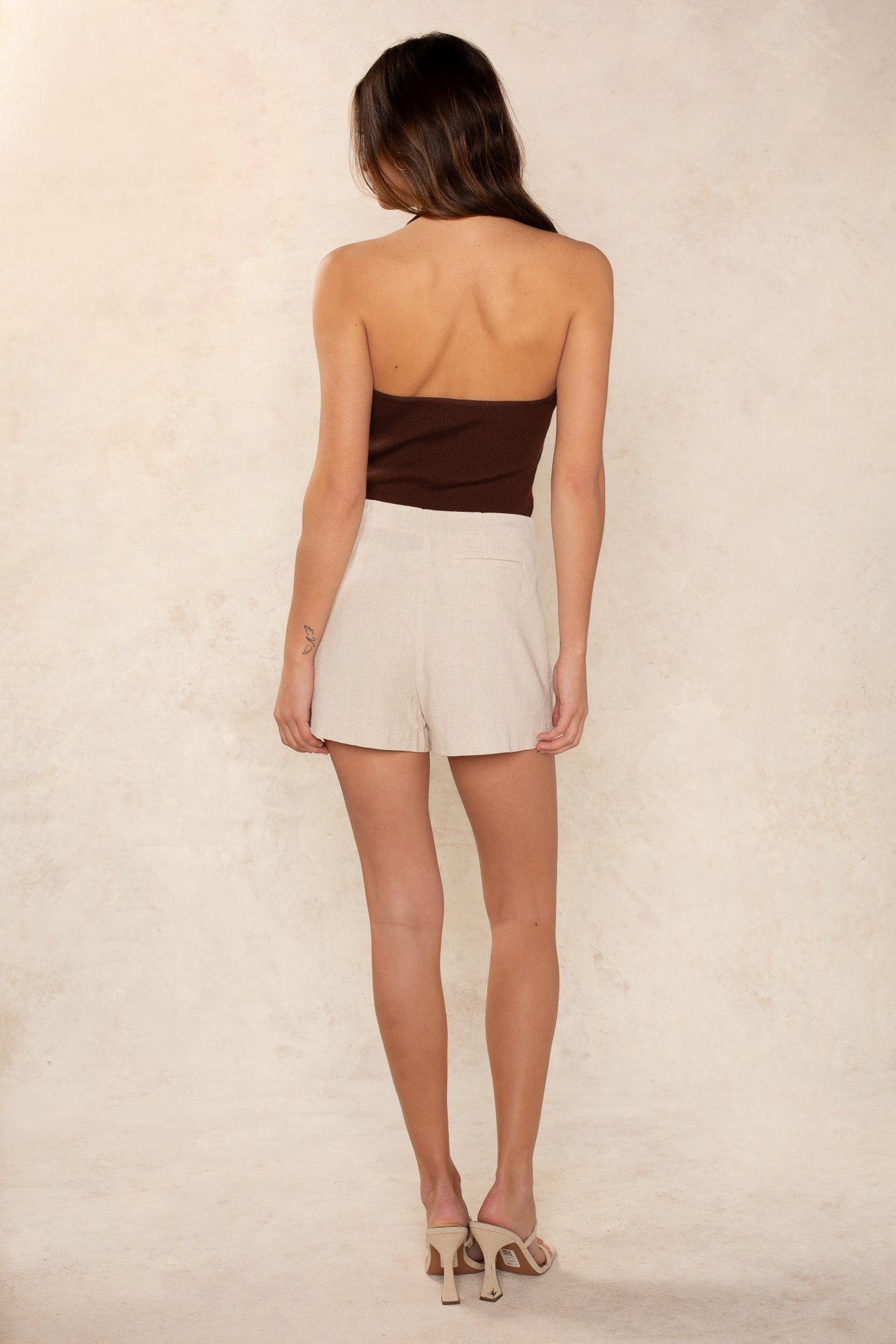 A woman with long brown hair stands facing away, wearing the Georgie Hailey Knit Halter, beige high-waisted shorts, and beige heeled sandals against a light textured background.
