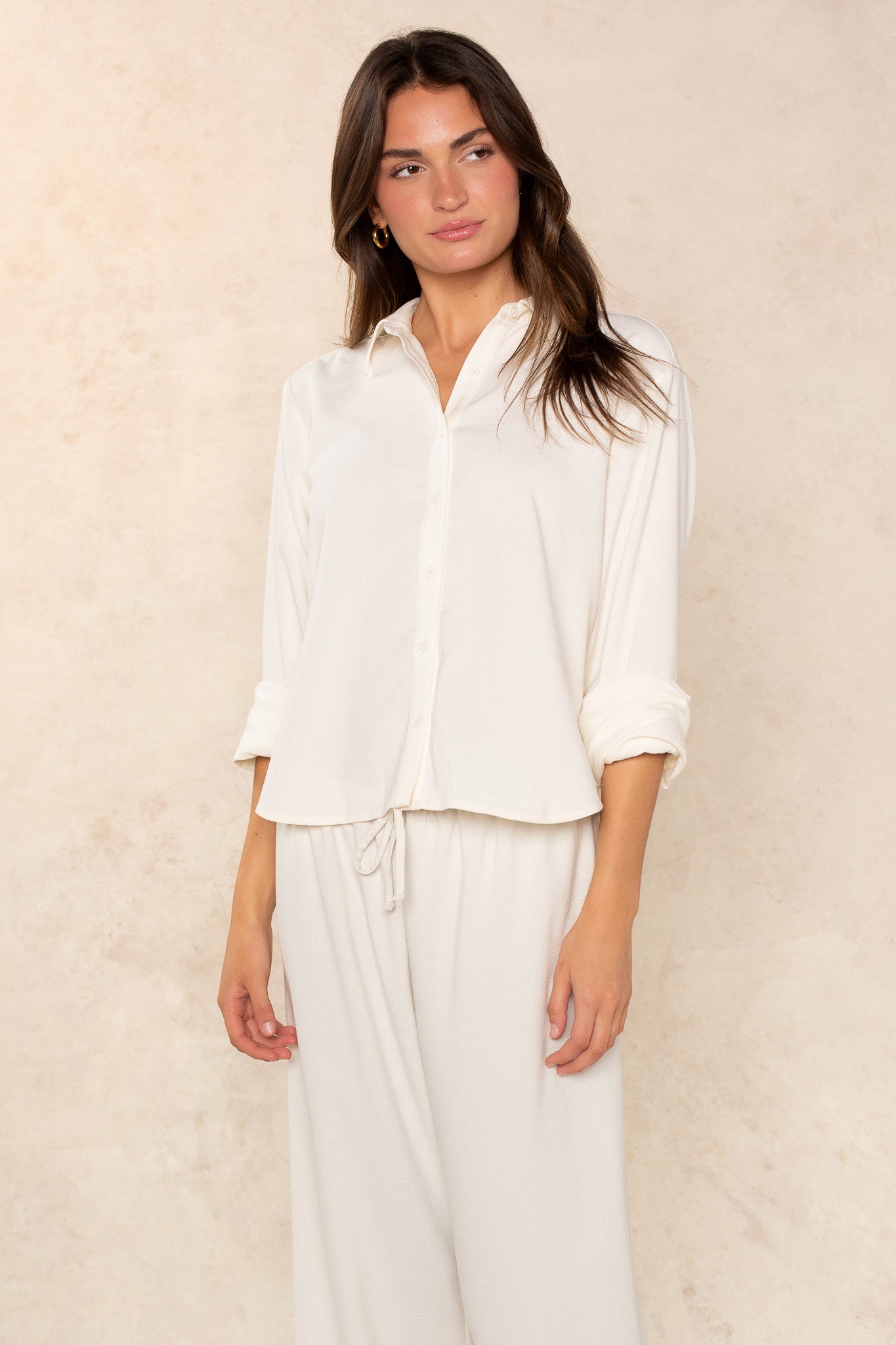 A woman wears the Georgie Seaside Blouse in white with sleeves rolled up and matching drawstring pants—timeless essentials—while standing against a light beige textured background.
