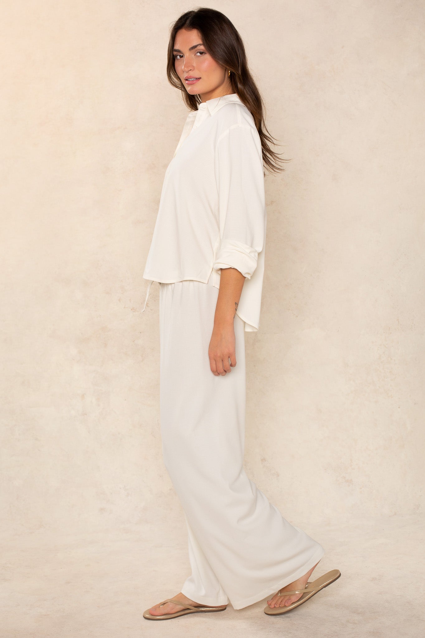 A woman with brown hair down stands sideways against a beige background, wearing the Georgie Seaside Blouse—an easy, long-sleeve white top paired with matching wide-leg pants. She looks relaxed and neutral; both pieces are crafted from quality fabric.