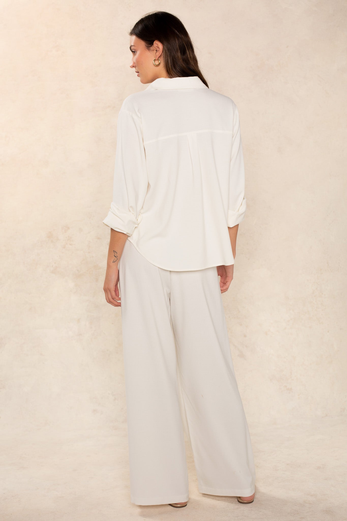 A woman with long brown hair poses against a light beige background, wearing Georgie’s Seaside Blouse—a wardrobe essential featuring a loose fit and long sleeves, paired with wide-leg trousers crafted from quality fabric.