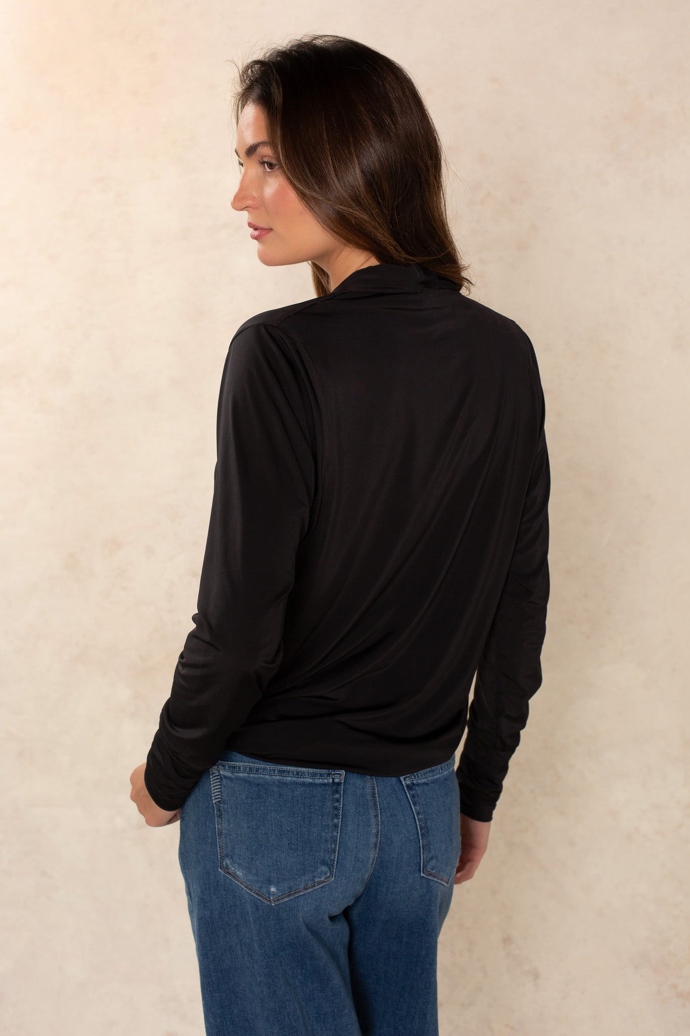 A woman with long brown hair wears the Georgie Gina Wrap Long Sleeve top in black and blue jeans, standing with her back turned and head slightly to the side against a light beige background.