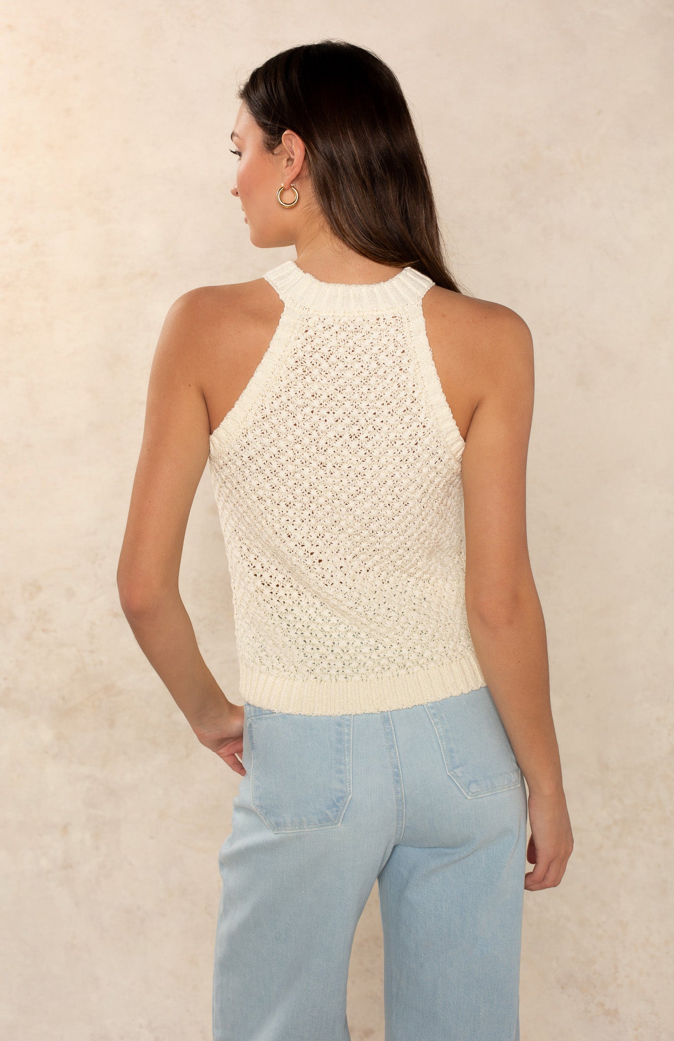 A woman with long brown hair wears the Georgie Ari Sleeveless Sweater, a cream knit racerback top, paired with light blue high-waisted jeans against a light beige textured background—an effortlessly timeless look.