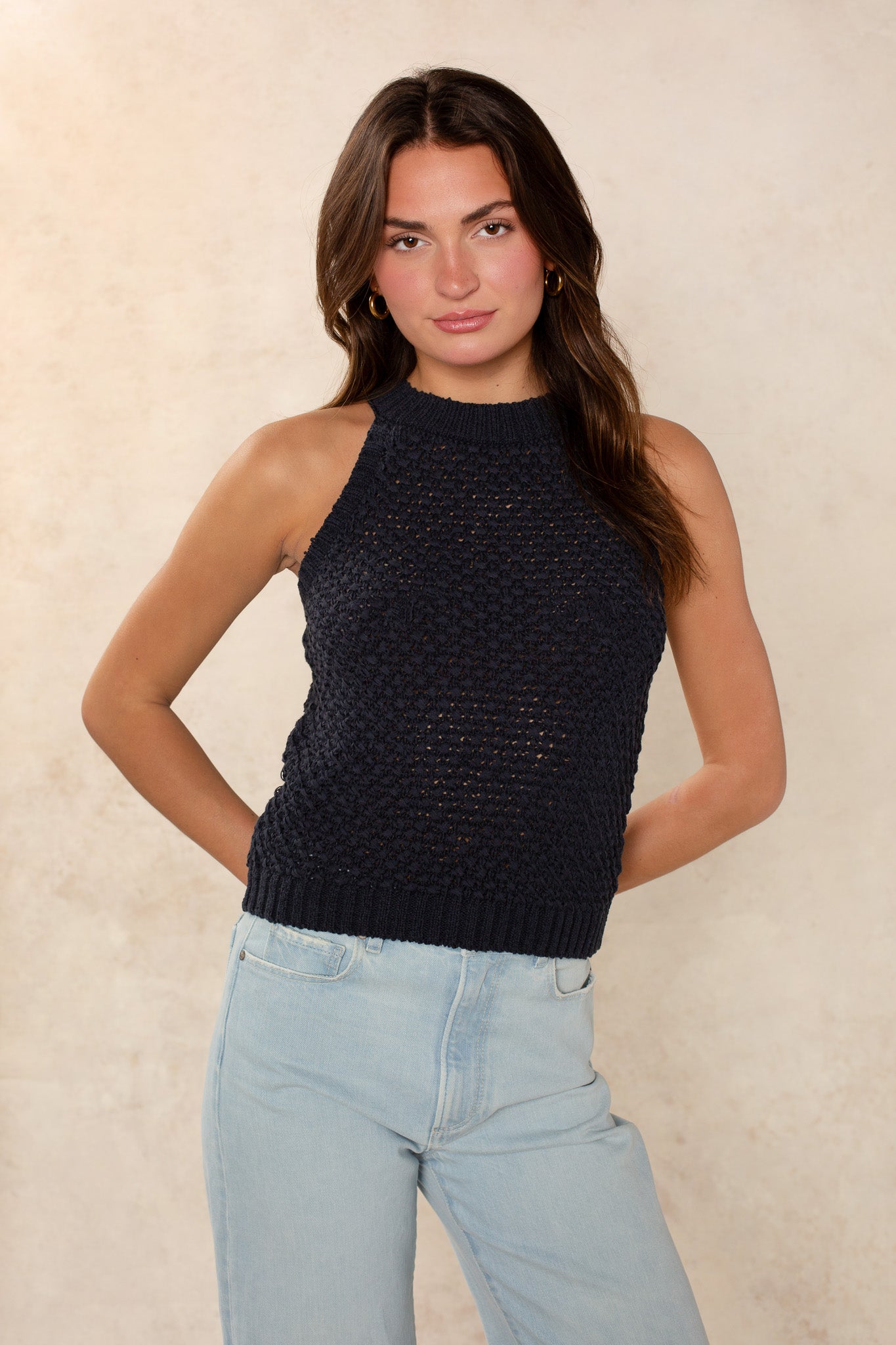 A woman with long brown hair models the Georgie Ari Sleeveless Sweater—a dark navy knit top—paired with light blue jeans, standing against a beige background with her hands behind her back.
