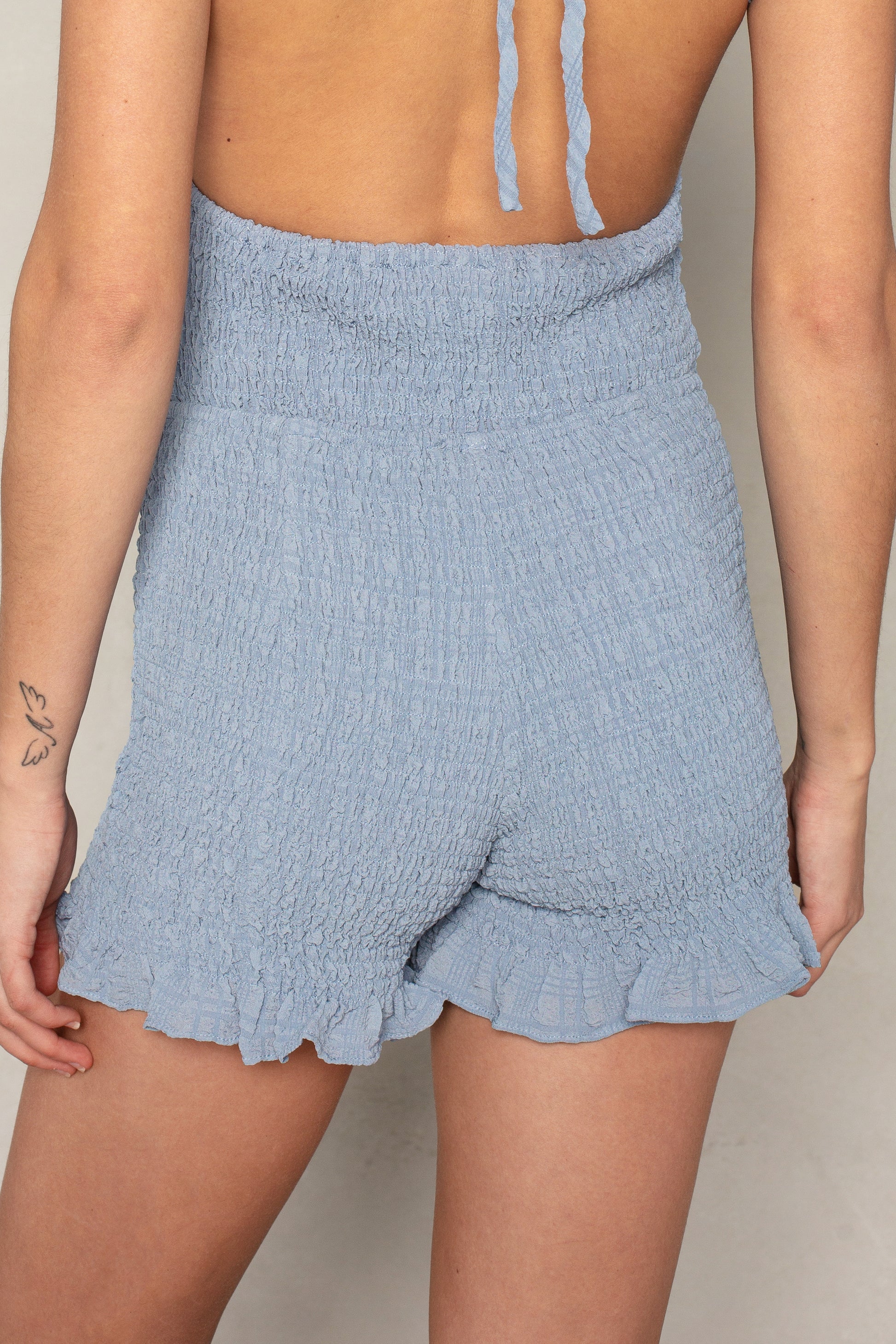 A person wearing the Isla x Tate Betty Smocked Short, a light blue textured strapless romper with mini shorts and ruffle hem, stands facing away. A small butterfly tattoo is visible on their left forearm.