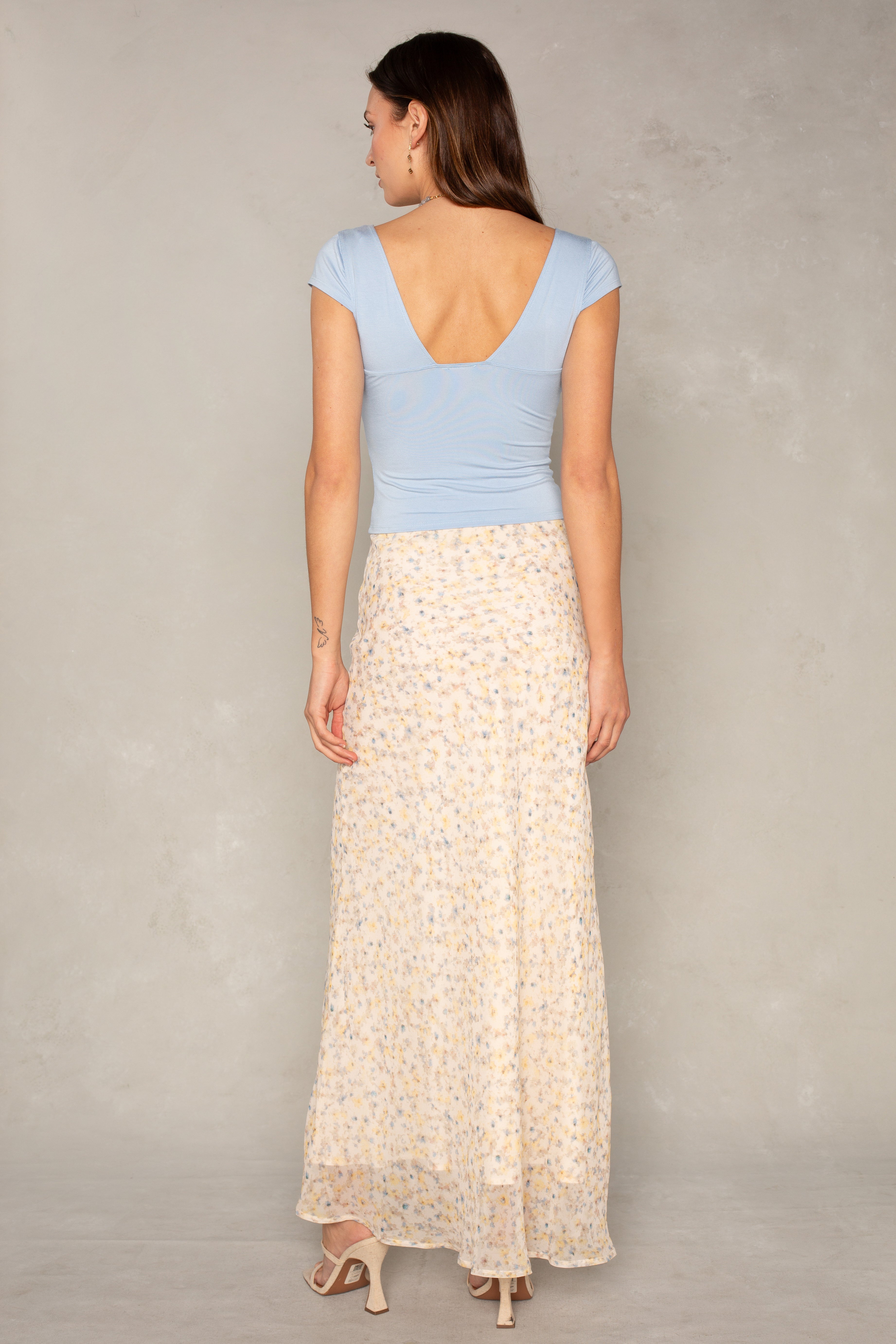 A woman stands with her back to the camera, wearing a light blue cap-sleeve top featuring a deep V-back, paired with the South Moon Under Mollie Floral Midi Skirt—a long, fit-and-flare chiffon skirt in a floral print—and heeled sandals. The backdrop is light gray.