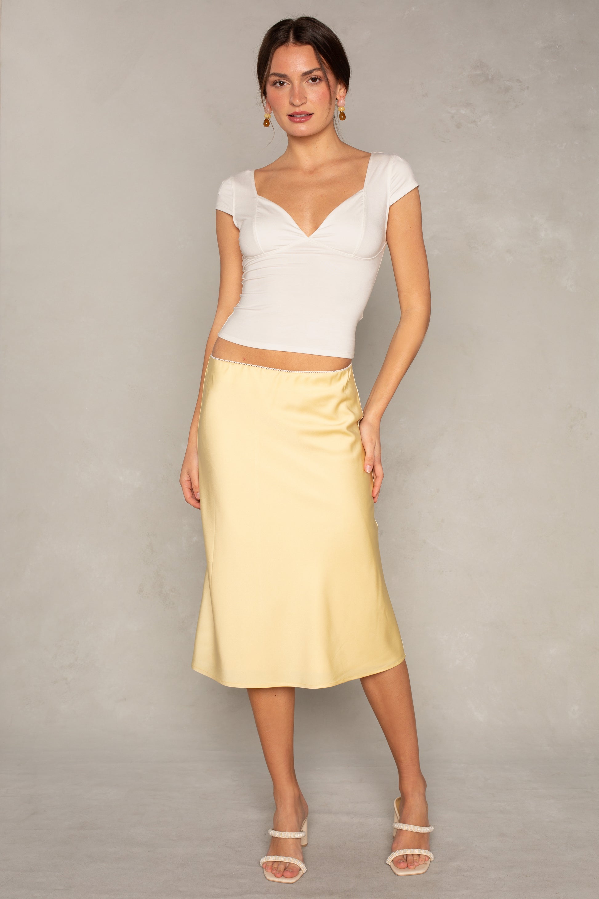 Against a light gray background, a woman wears the Isla x Tate Emme Satin Skirt in light yellow with a fitted and flared silhouette, styled with a white cap-sleeve top, white heeled sandals, and drop earrings. She poses gracefully, one hand slightly raised.