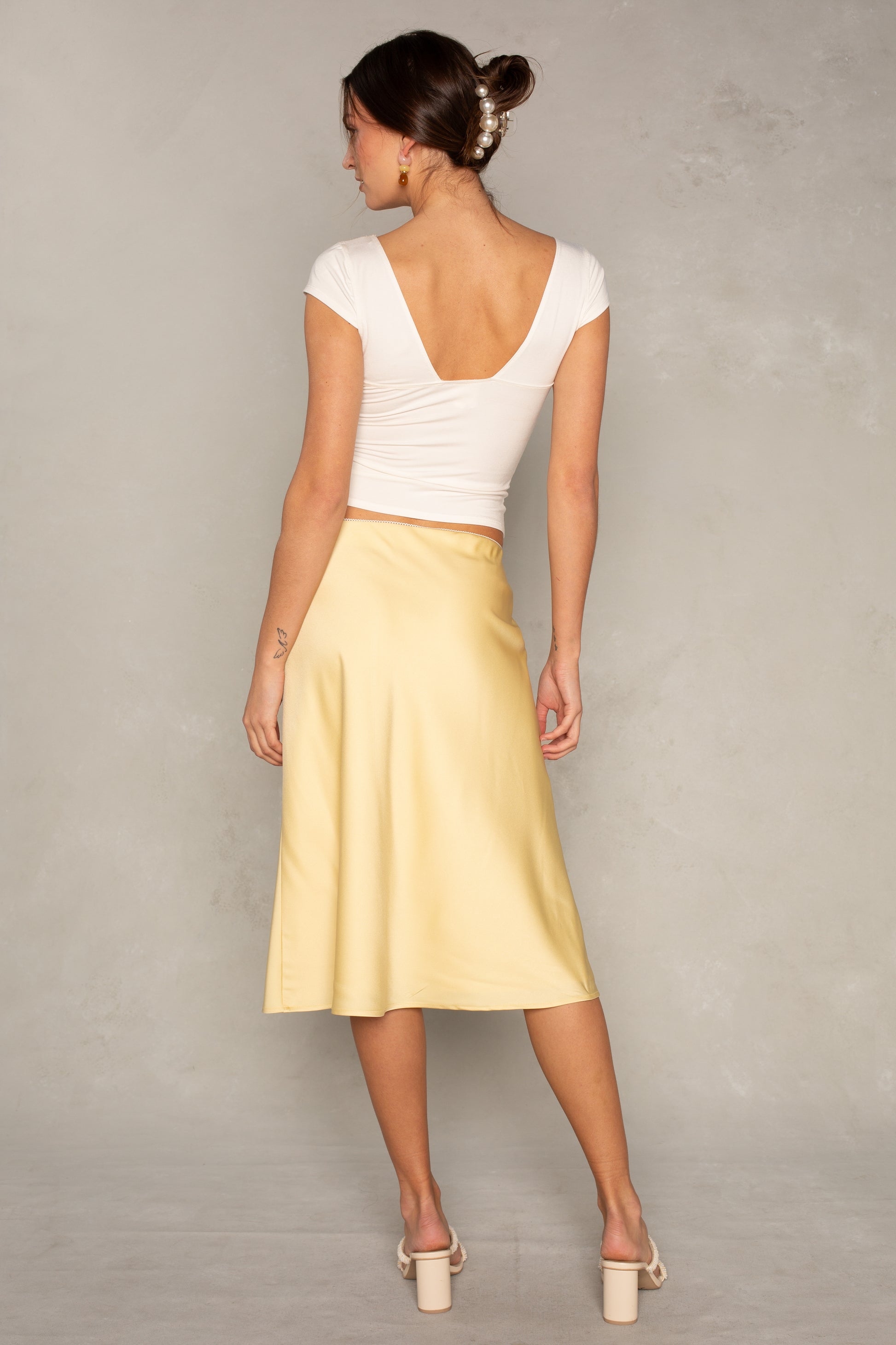 A woman, with her hair in a low bun and wearing heeled sandals, models the Isla x Tate Emme Satin Skirt in light yellow with a knee-length fit, paired with a fitted white cap-sleeve top featuring a deep V-back.