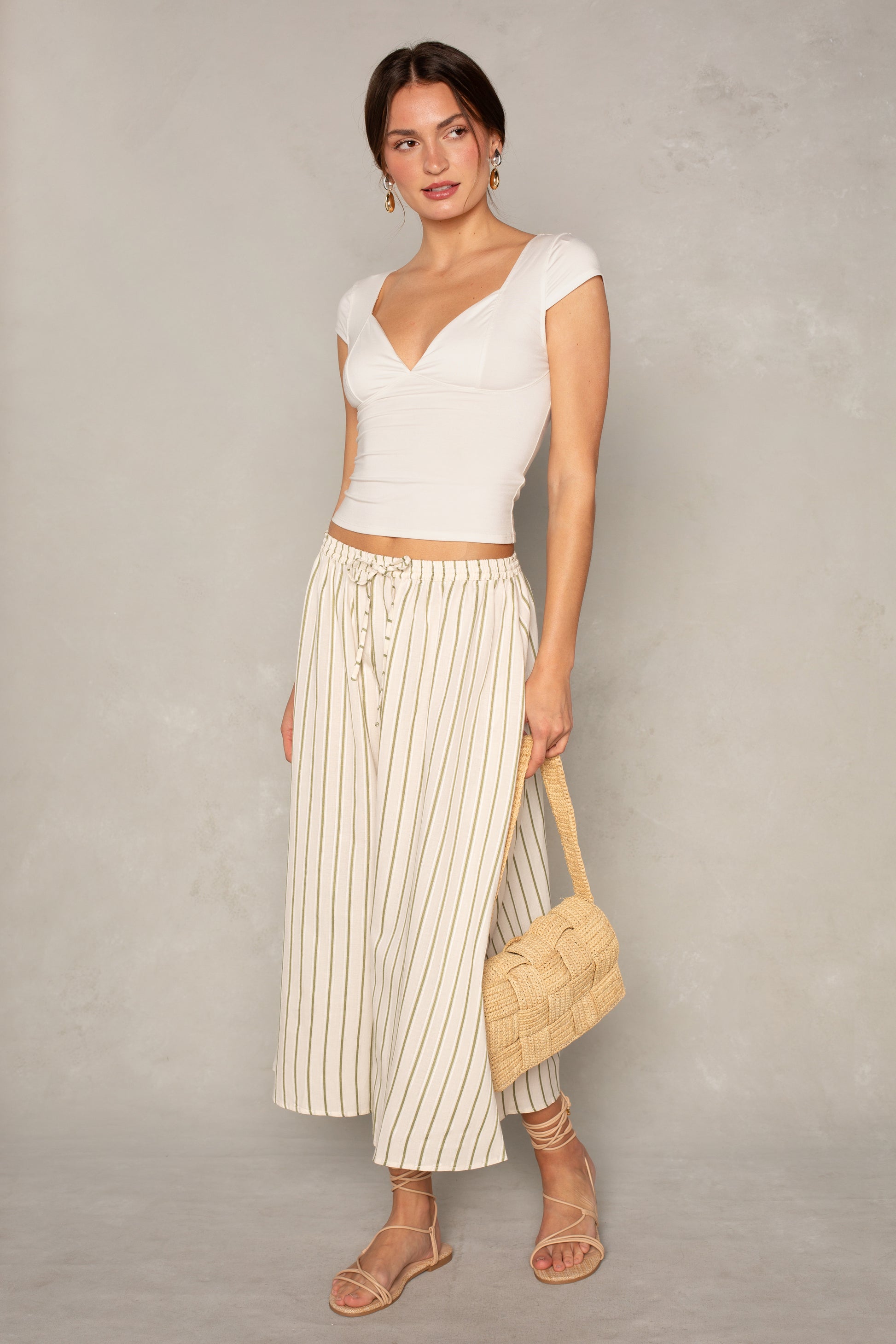 A woman wearing the Isla x Tate Grace Stripe Midi Skirt—cream with thin vertical stripes and an elastic waist—paired with a white fitted top, woven handbag, and strappy sandals stands against a light, neutral background.