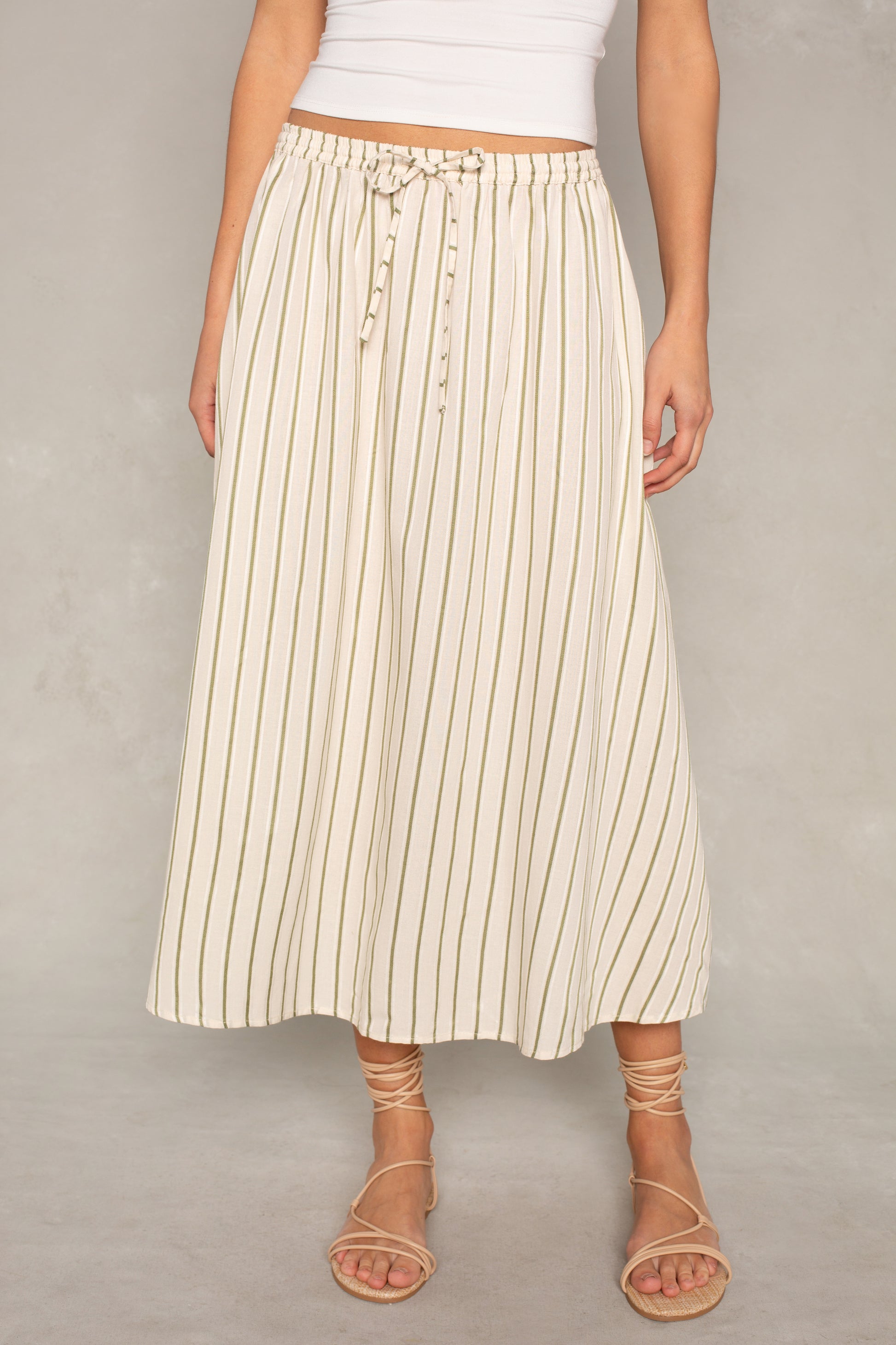 Someone wearing the Isla x Tate Grace Stripe Midi Skirt—a white, high-waisted, A-line midi skirt with thin vertical beige stripes and an elastic waist—paired with beige strappy sandals, stands against a plain background.