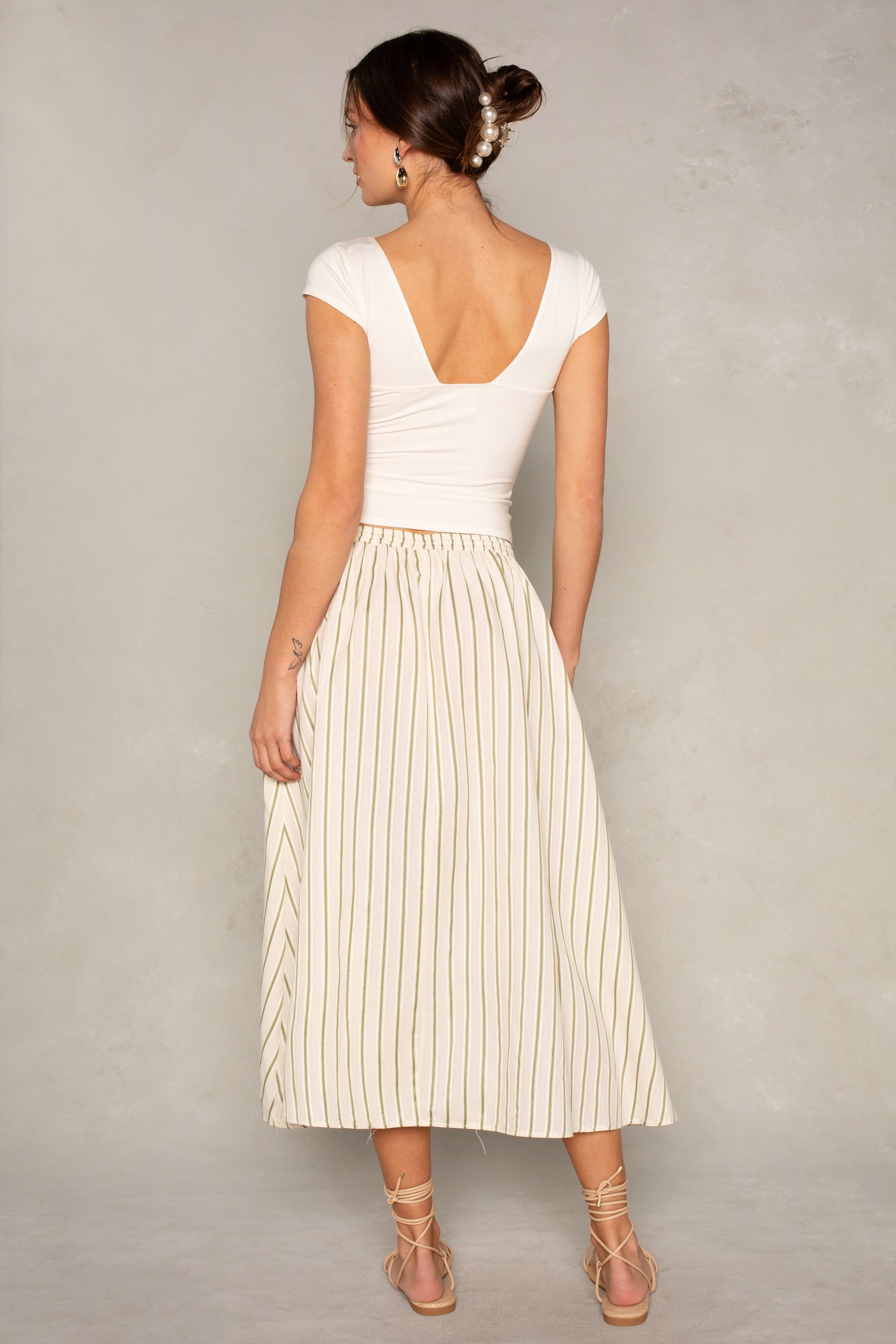 A woman faces away, wearing the Isla x Tate Grace Stripe Midi Skirt with thin vertical stripes and an elastic waist, paired with a white cap-sleeve deep V-back top, her hair in a low bun with clips, and strappy nude sandals.