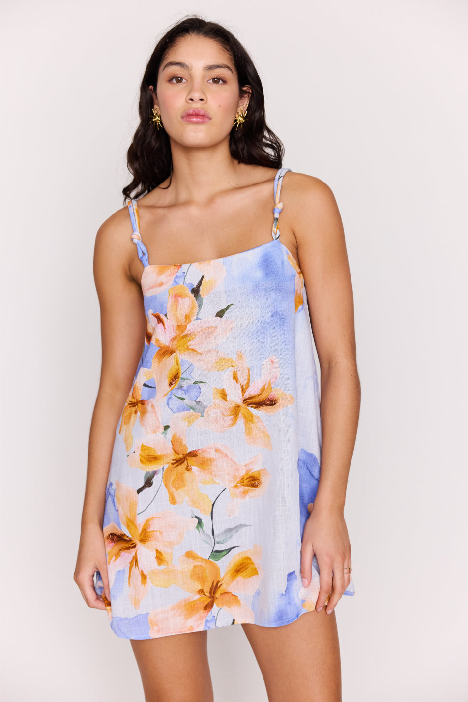A woman in the Minkpink Sunspell Knot Strap Mini Dress—light blue with orange and yellow floral prints—stands against a plain light background, gazing at the camera with a neutral expression.
