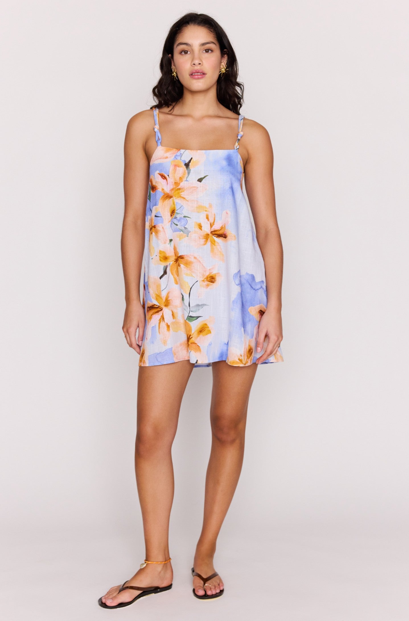 A woman with dark hair faces forward, wearing the Minkpink Sunspell Knot Strap Mini Dress in watercolour floral print with blue and orange hues. She is barefoot except for strappy sandals, posing against a plain light background.