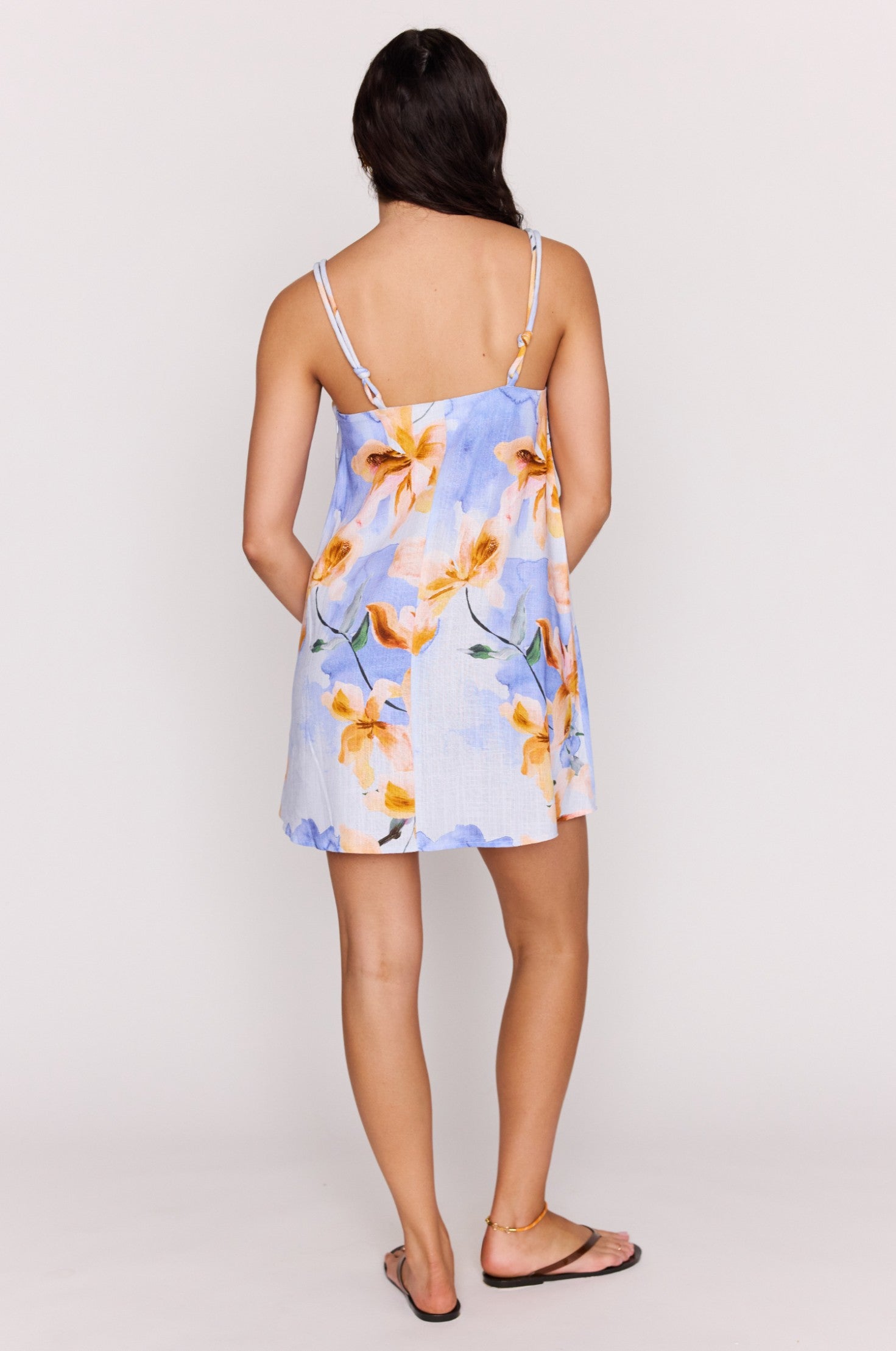A woman faces away, wearing the Minkpink Sunspell Knot Strap Mini Dress—a sleeveless, light blue mini dress with yellow and orange floral print—and black flip-flops against a plain light background.