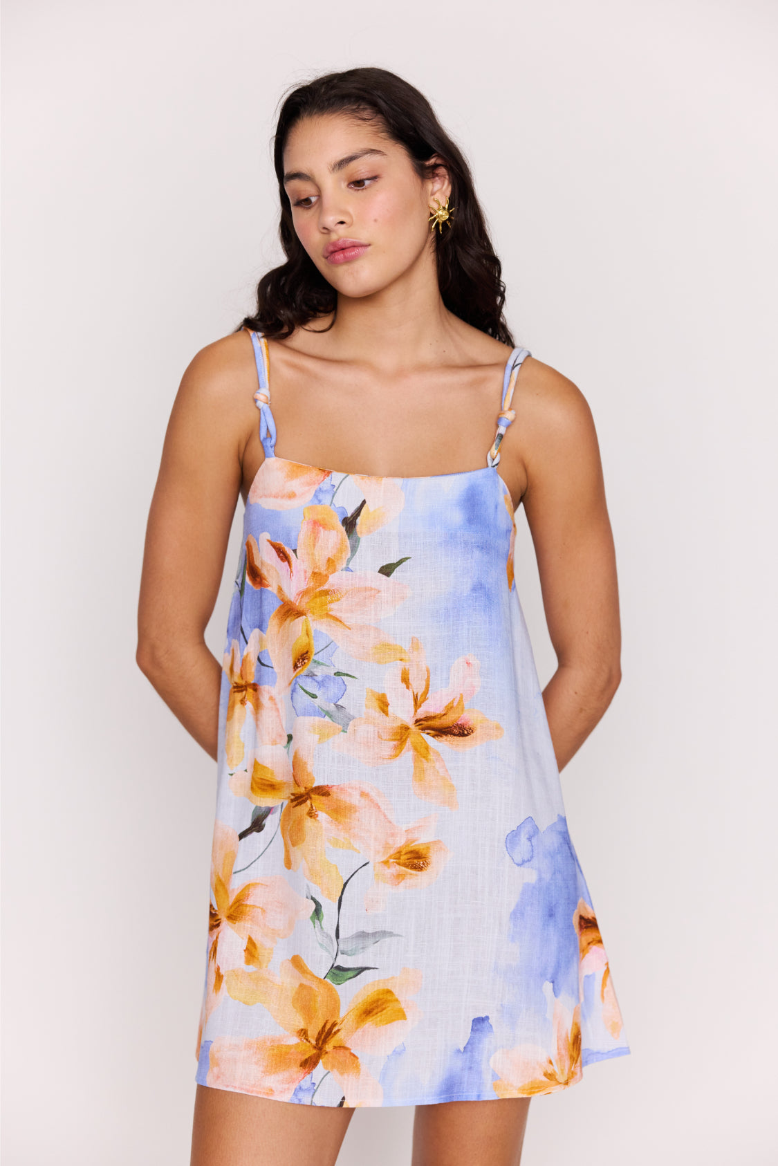 A woman wearing a sleeveless Sunspell Knot Strap Mini Dress by Minkpink in Blue Multi stands against a light background, looking slightly down with her hands behind her back.