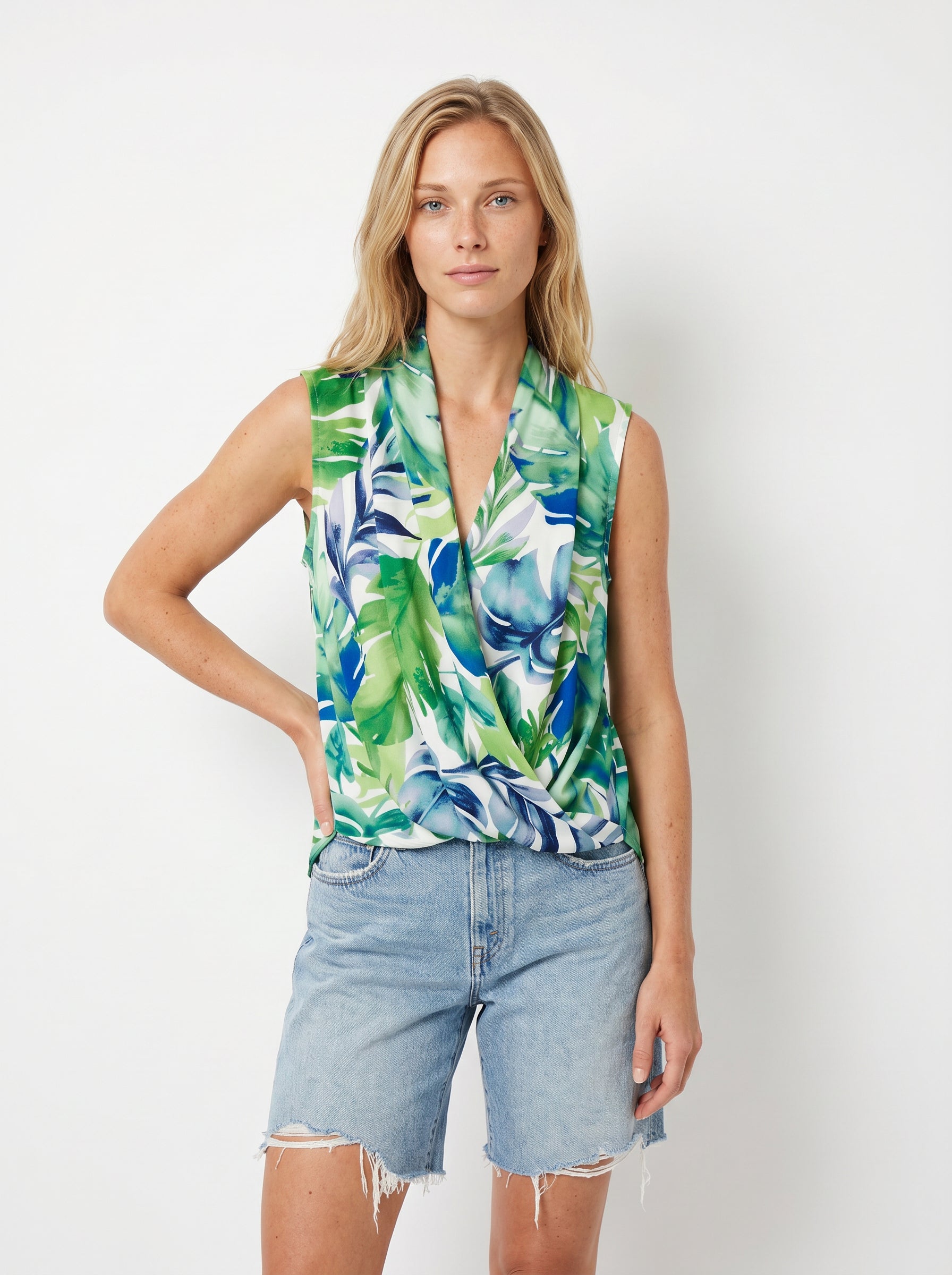 A person with long blonde hair wears the Georgie Gwen Sleeveless Wrap Top in GREEN MULTI and light denim cut-off shorts, standing against a plain white background.