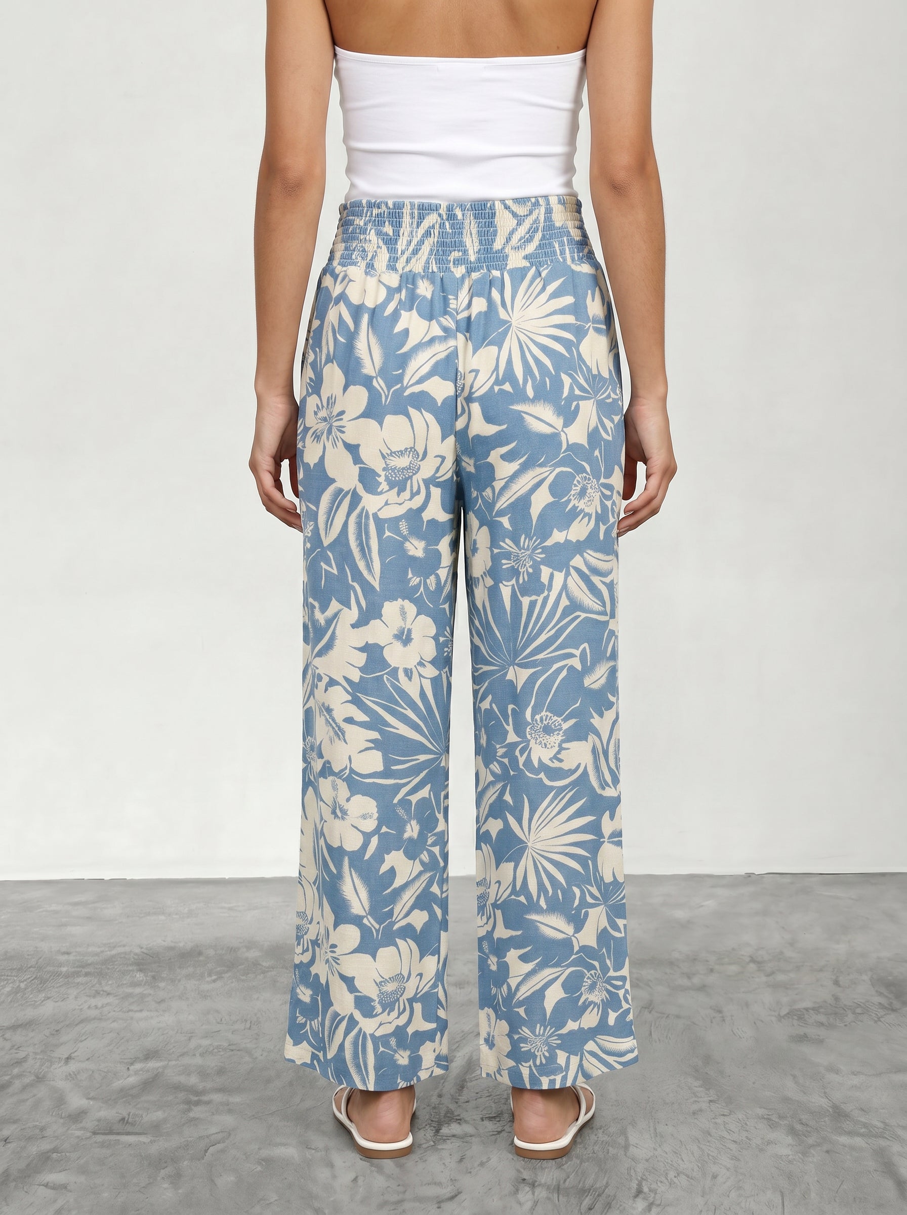 A woman stands on a gray floor, shown from behind against a light background, wearing Isla's Hattie Beach Pant in a blue and white floral print, paired with white heeled sandals and a white tube top.