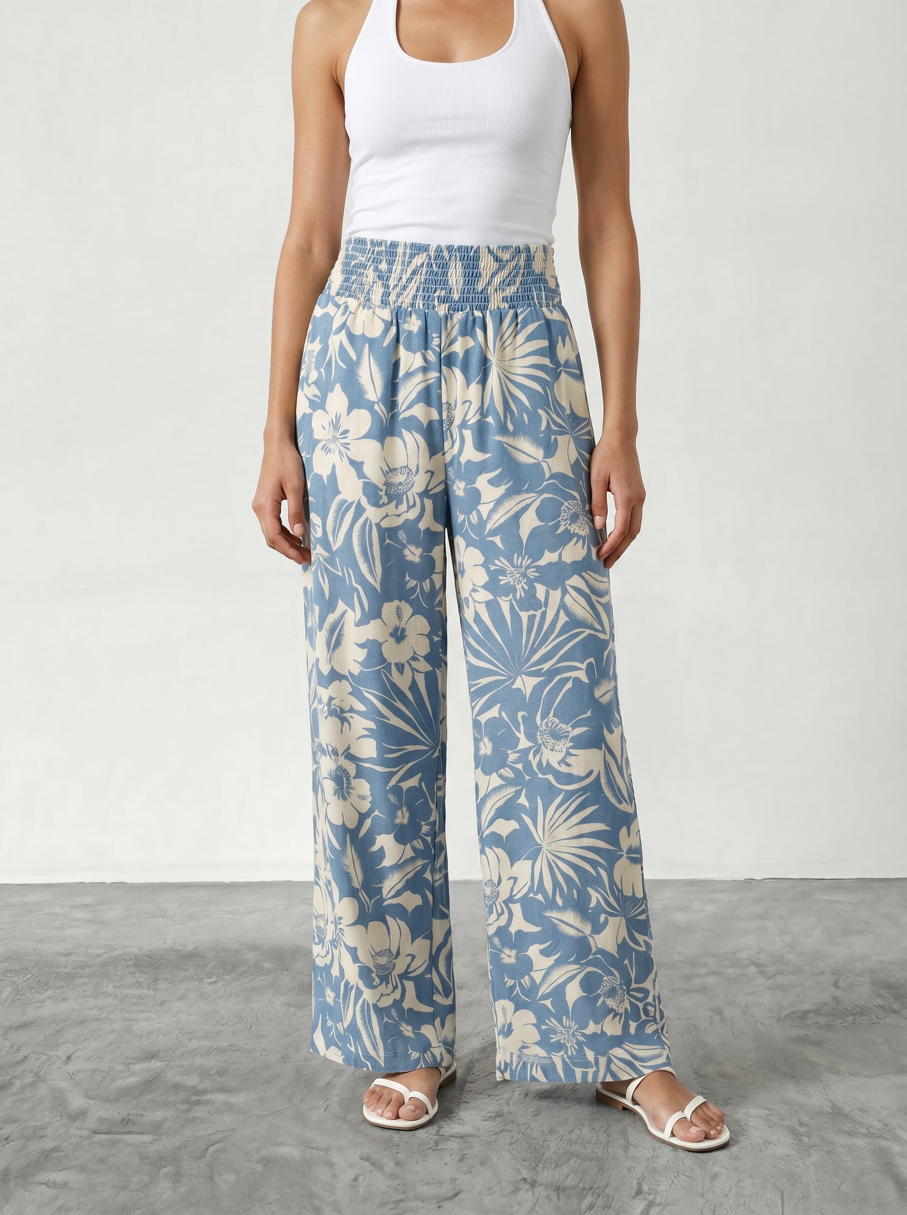 A person wearing an Isla Hattie Beach Pant—blue and white floral wide-leg style with an elastic waistband—a white tank top, and white sandals stands on a gray floor against a plain light background.