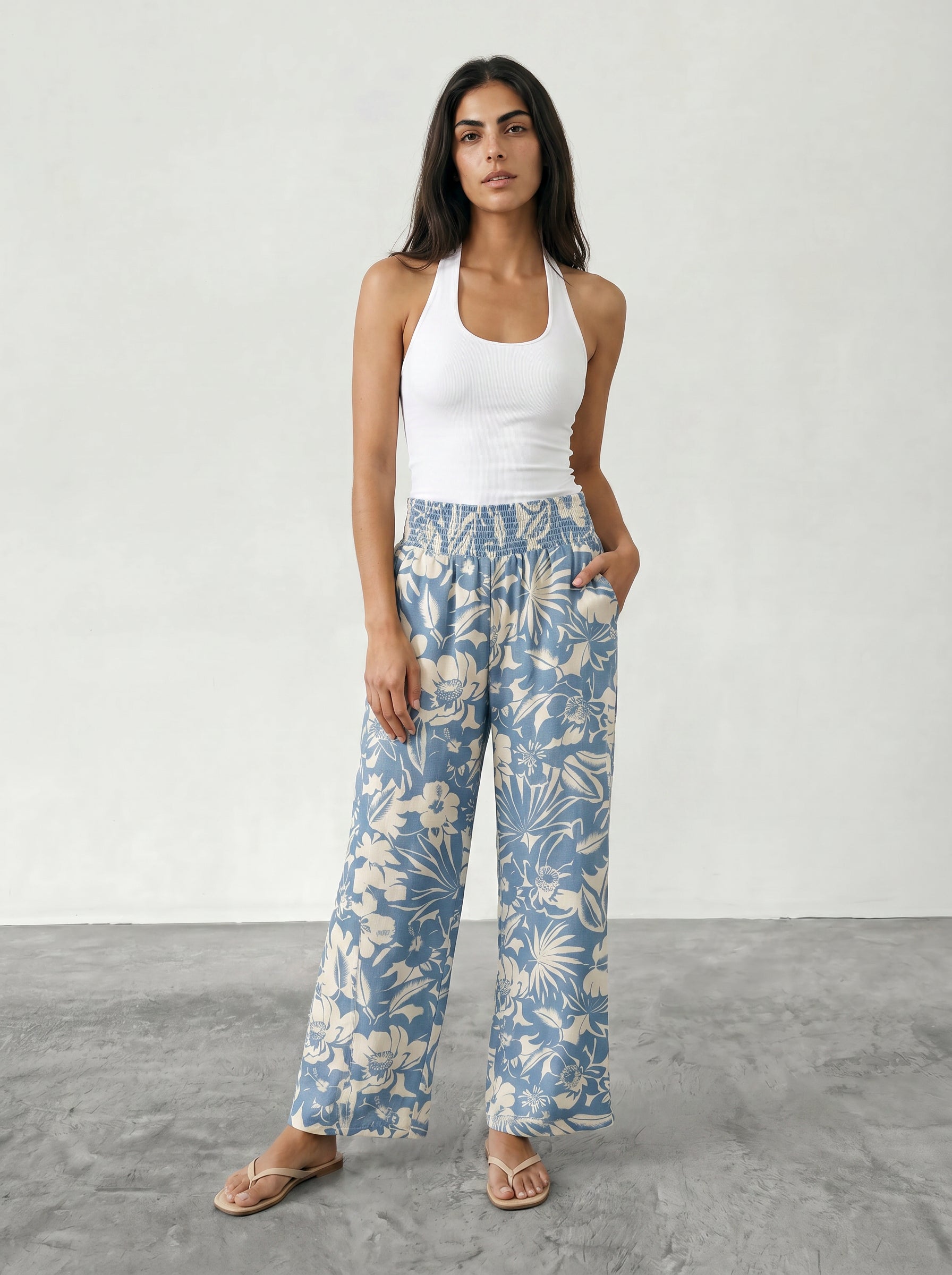 A woman with long dark hair stands on a gray floor, looking at the camera with one hand in her pocket. She wears beige sandals, a white halter top, and Isla’s Hattie Beach Pant—blue wide-leg pants with a white floral pattern.