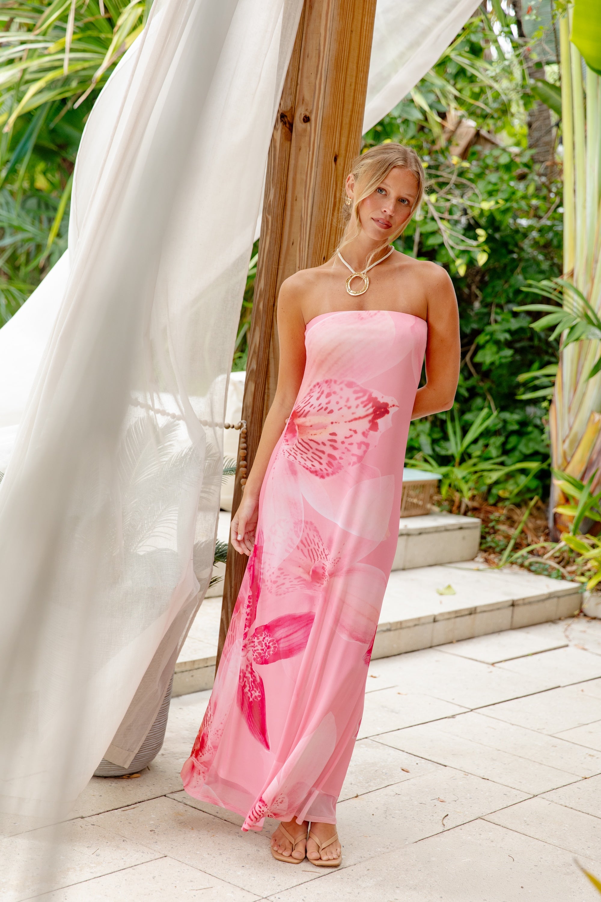 A woman poses outdoors by a wooden pillar wearing the Isla Flora Tube Mesh Maxi Dress in Pink Multi, featuring large floral patterns. She accessorizes with a statement necklace, with green foliage and white drapes in the background.