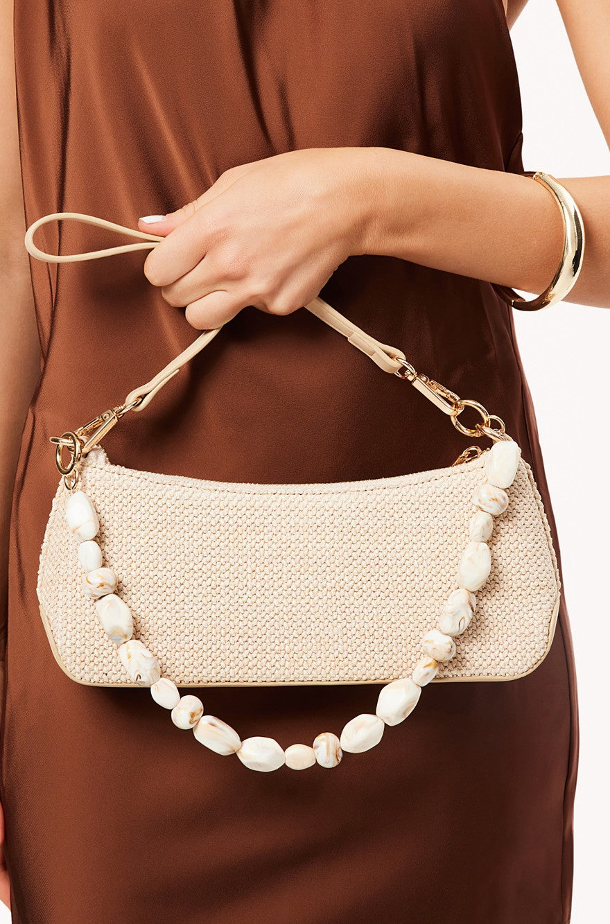 A person in a brown dress holds the Jamie Shoulder Bag by Billini, a cream-colored rectangle bag with gem embellishments, a beaded handle, and gold hardware. They also wear a gold bangle on their wrist.