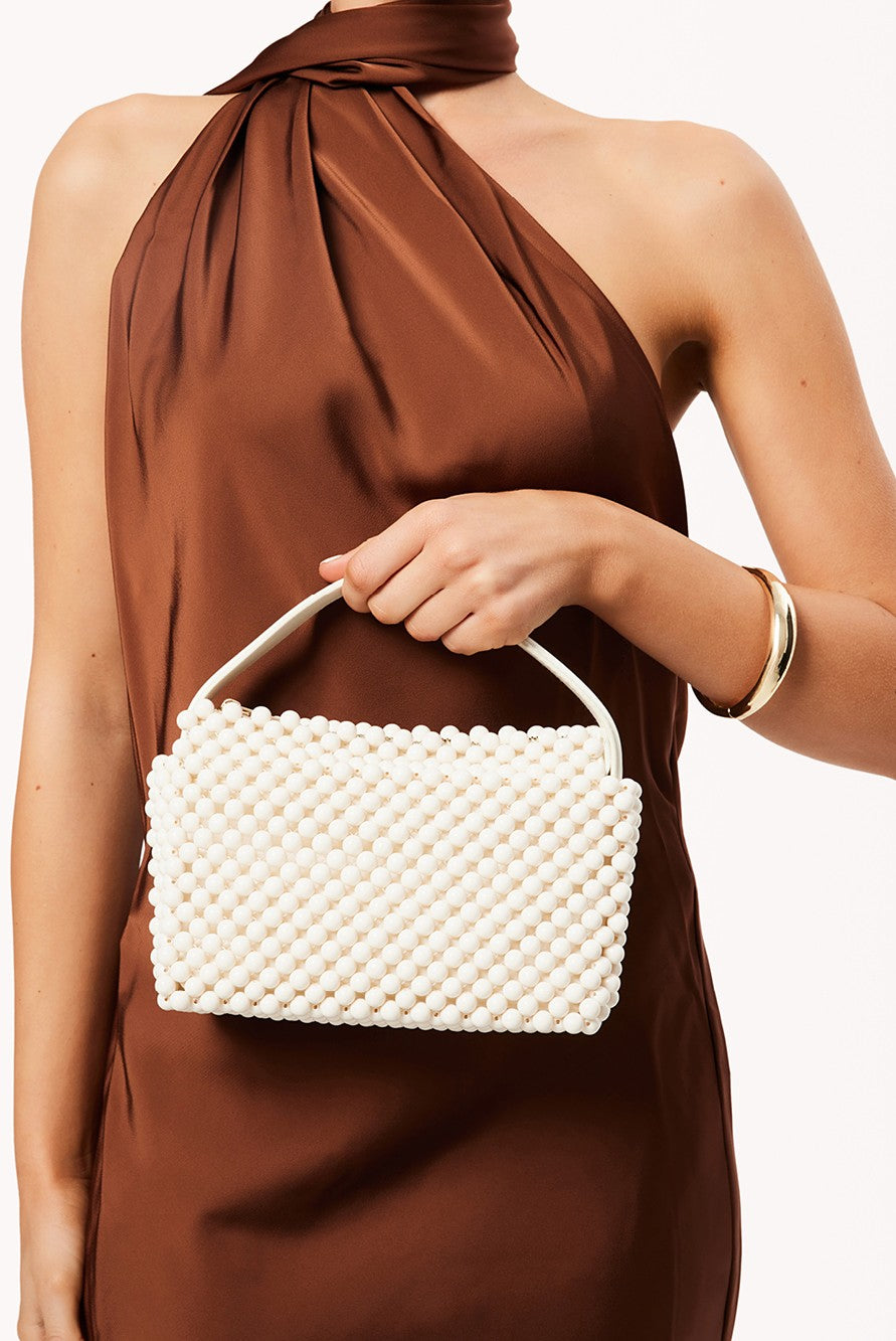 A woman in a sleeveless brown dress holds the Billini Jeanie Handle Bag in ivory, featuring beaded detailing and a top handle, in front of her torso. She accessorizes with a gold bangle against a plain light background.