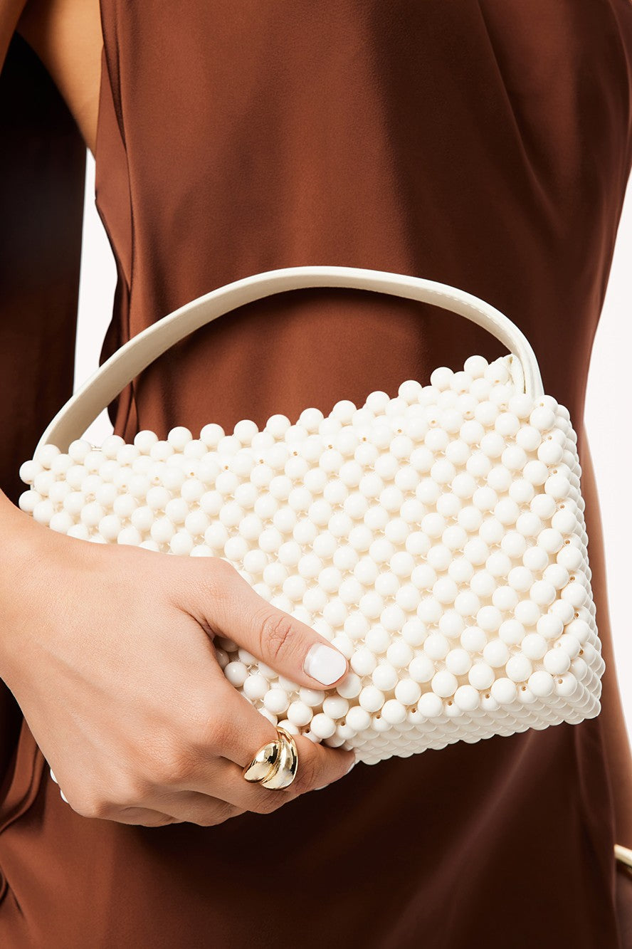 A person in a brown satin outfit holds the Billini Jeanie Handle Bag, a rectangular handbag with white beaded detailing and a top zip closure, accessorized with neat nails and a chunky gold ring.