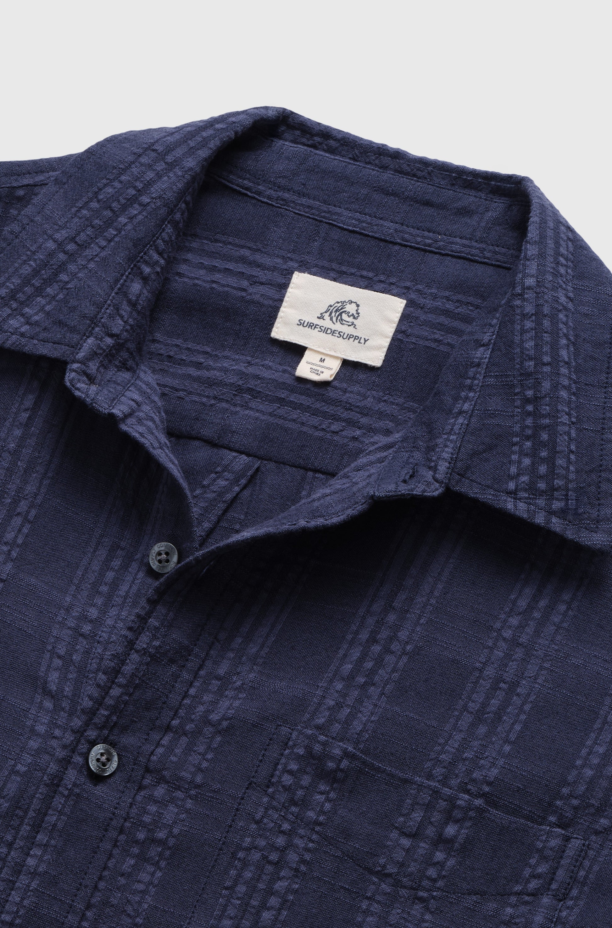 Close-up of the Joseph Straight Bottom Textured Shirt by Surfside Supply Co. in navy, featuring black buttons and a white label inside the collar. The cotton rayon blend shirt is laid flat on a light background.