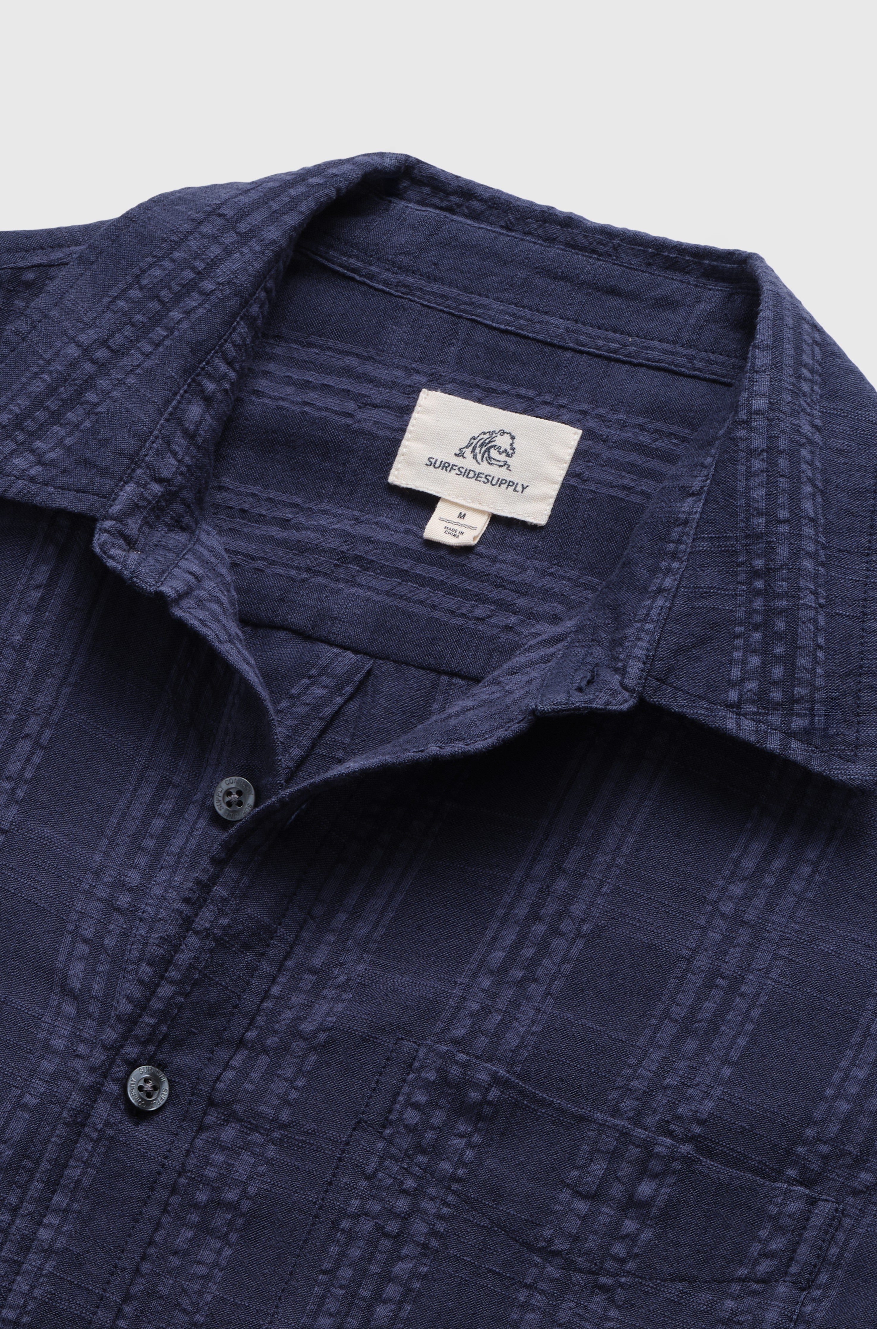 Close-up of the Joseph Straight Bottom Textured Shirt by Surfside Supply Co. in navy, featuring black buttons and a white label inside the collar. The cotton rayon blend shirt is laid flat on a light background.