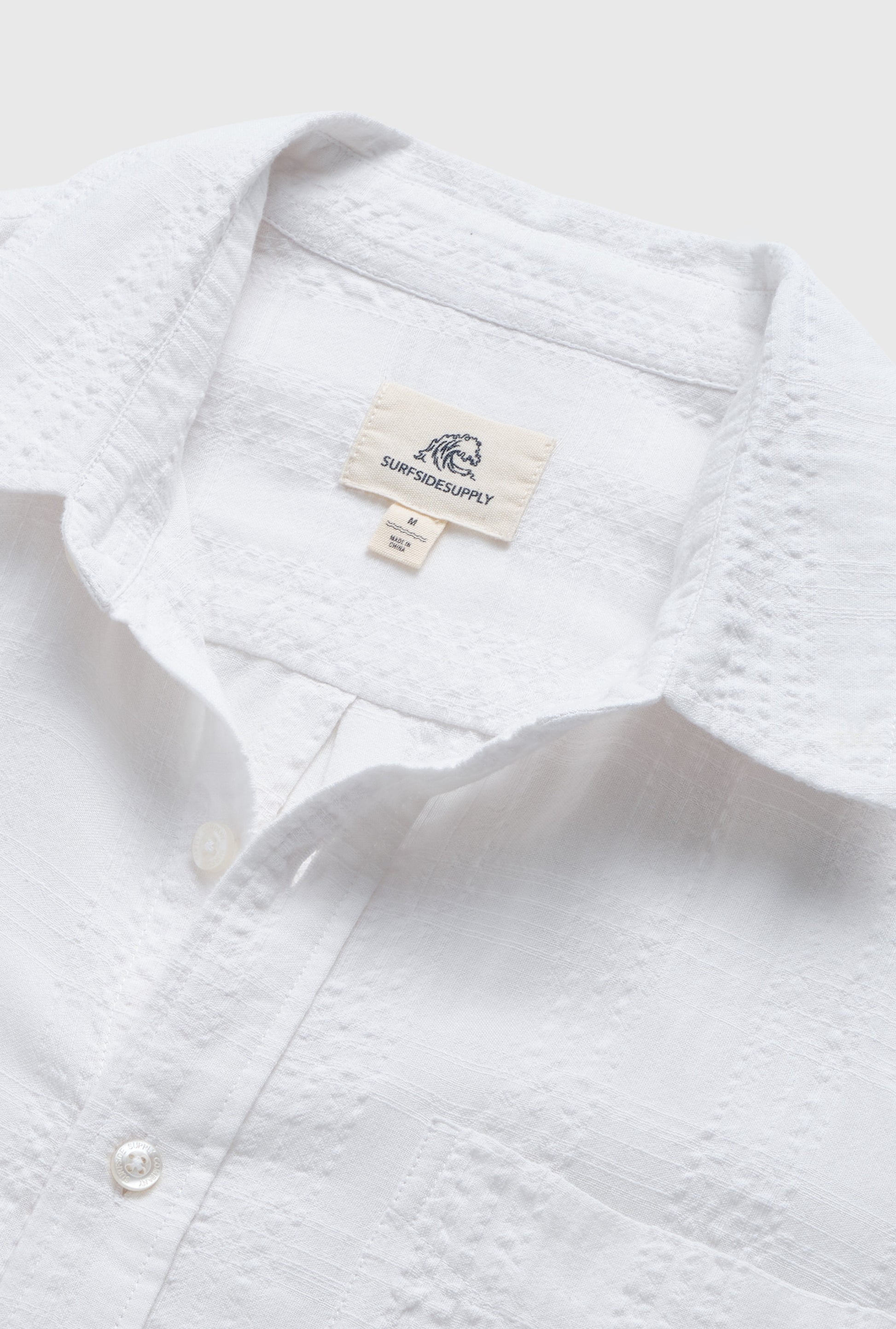 Close-up of the Joseph Straight Bottom Textured Shirt in white by Surfside Supply Co., featuring a pointed collar, regular fit, and soft cotton-rayon blend. The label with wave logo marks its medium size.