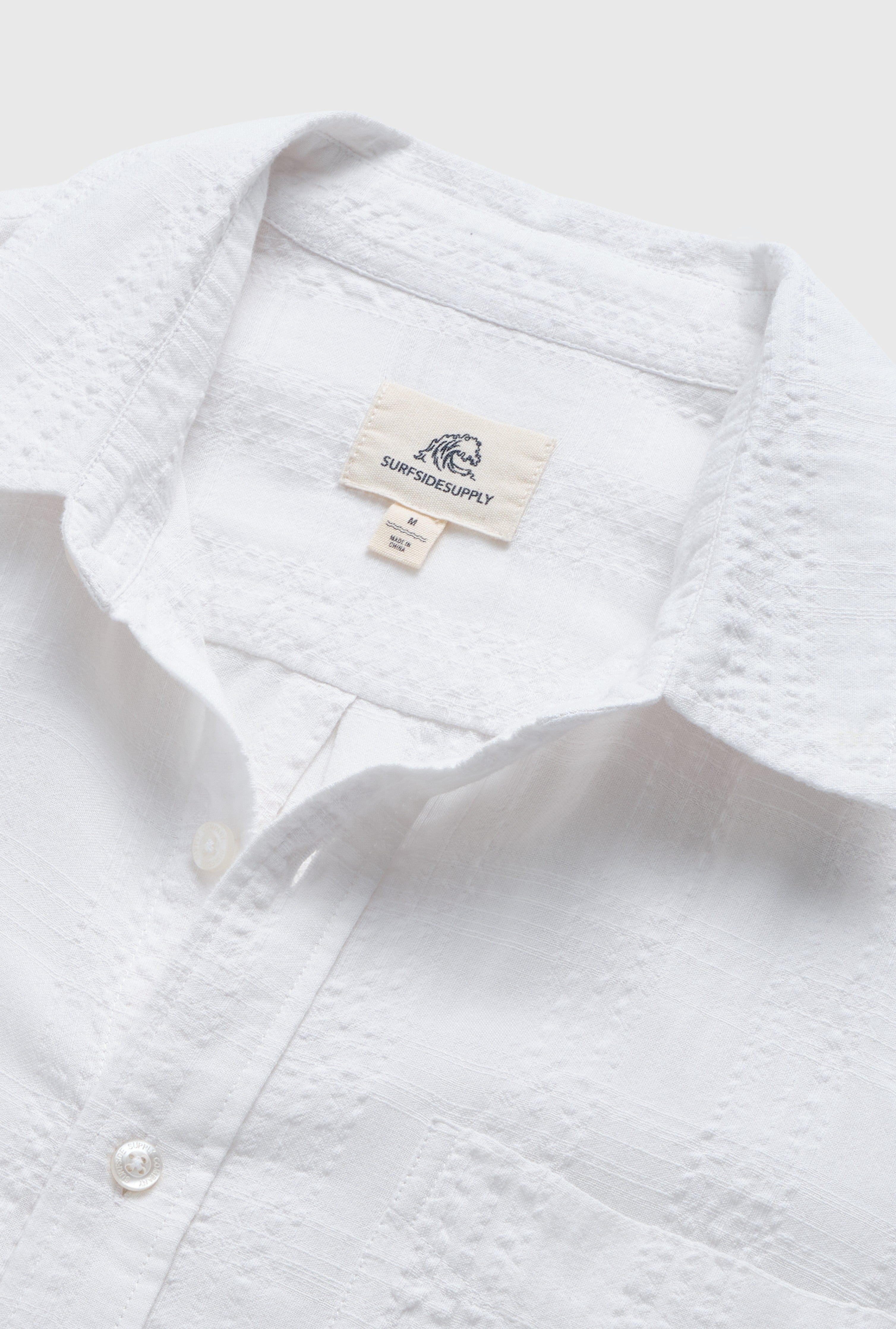Close-up of the Joseph Straight Bottom Textured Shirt in white by Surfside Supply Co., featuring a pointed collar, regular fit, and soft cotton-rayon blend. The label with wave logo marks its medium size.