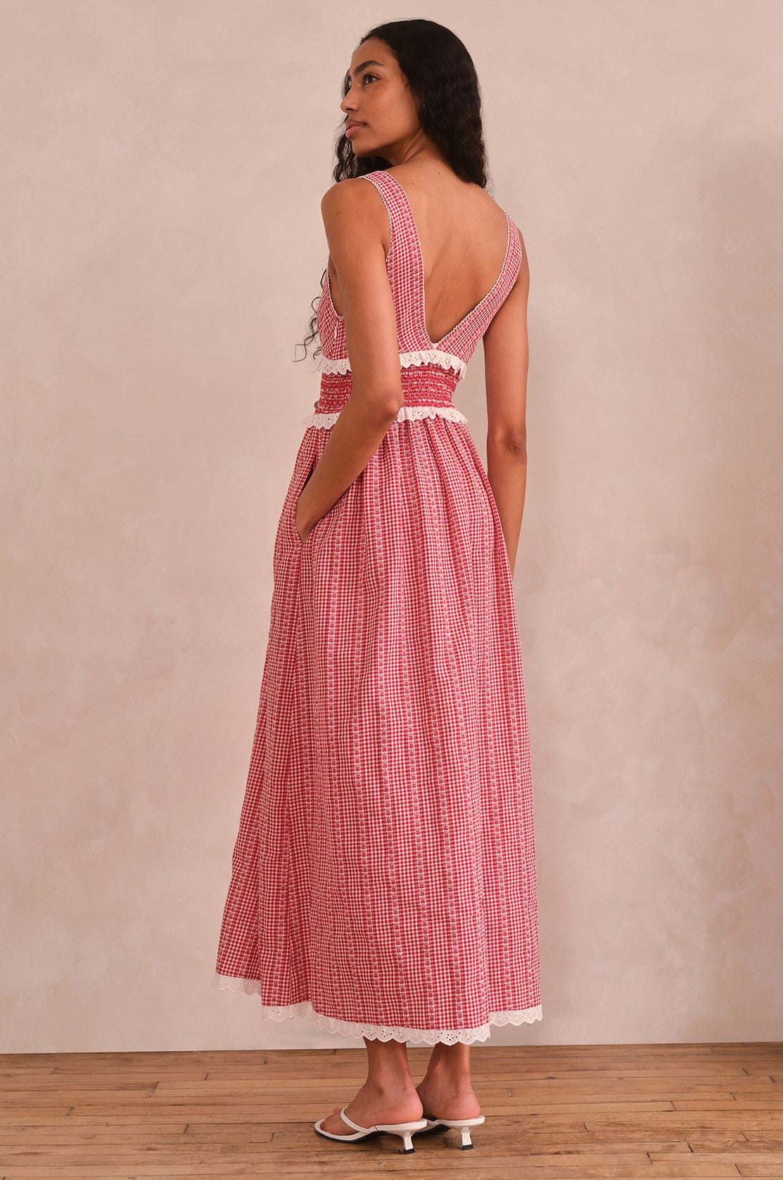A woman with long wavy hair stands on a wooden floor, facing away, wearing Saylor’s Karima Maxi Dress—a red and white floral gingham style with a smocked bodice, deep back, lace trim, pockets, ankle-length hem—and white sandals.