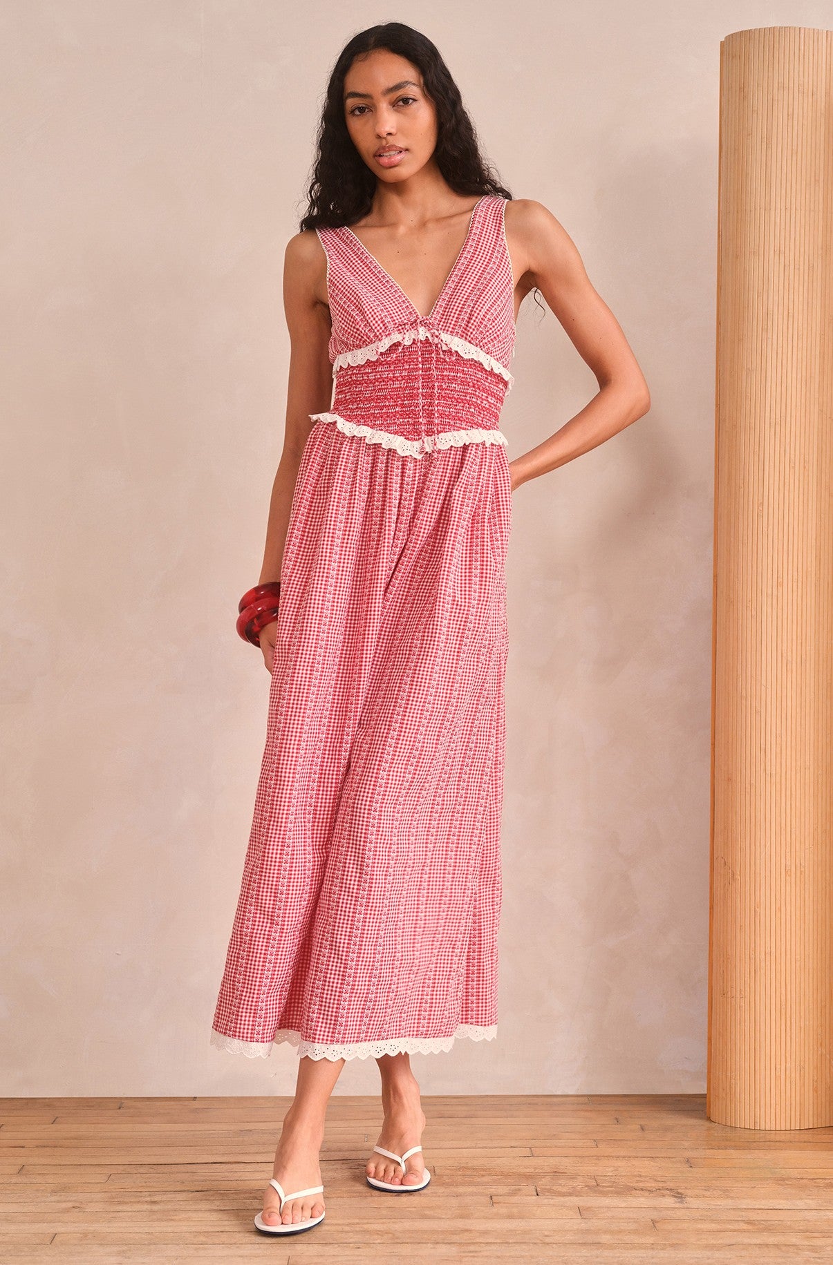 A woman poses indoors on wooden flooring by a column, wearing the Saylor Karima Maxi Dress—a sleeveless, red and white floral gingham maxi dress with lace trim, a smocked bodice, and a plunge v-neckline.