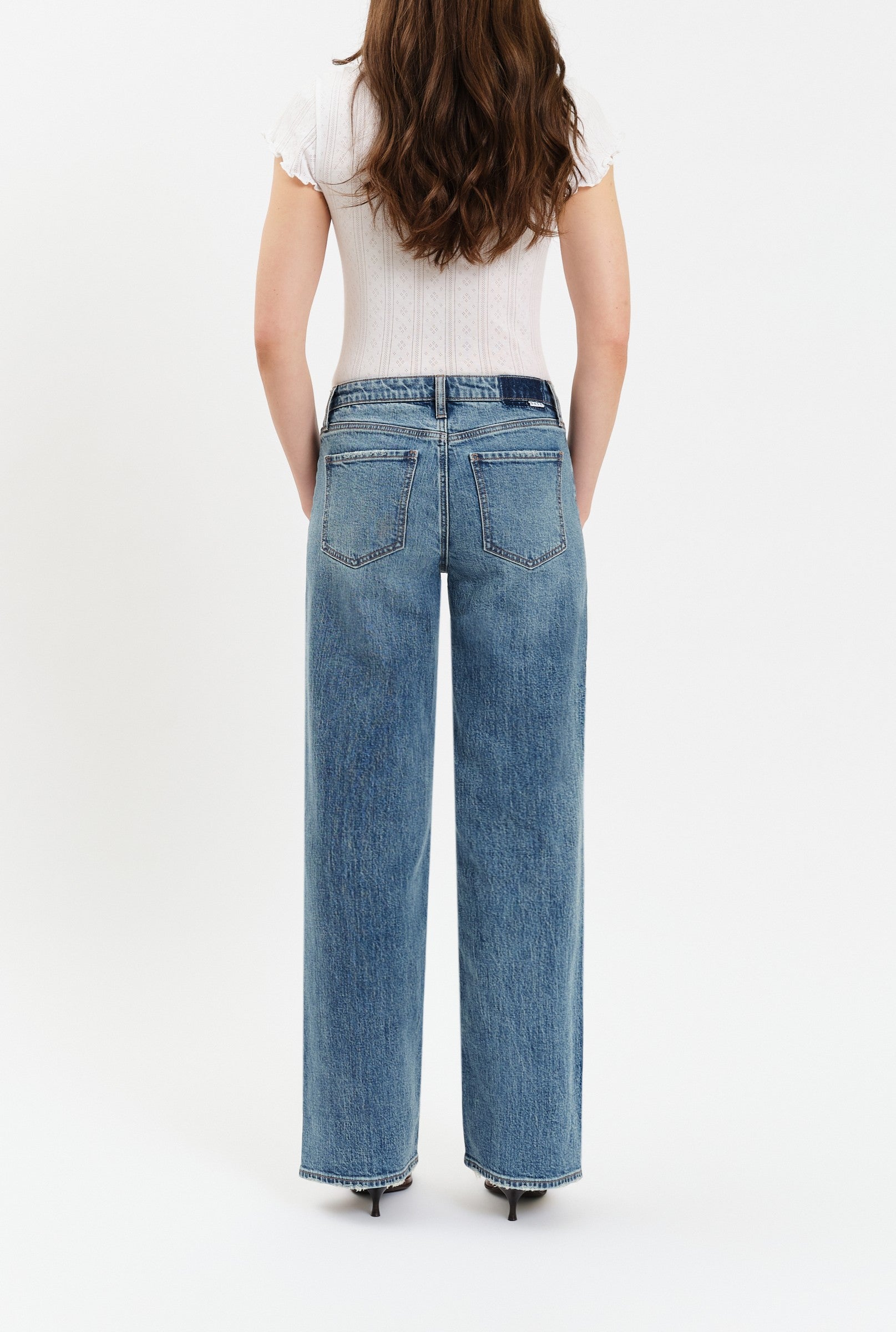 A woman stands with her back to the camera, wearing Daze’s The Keeper Low Rise In Heartbeat Vintage—high-waisted, wide-leg blue jeans made from comfort stretch denim with visible back pockets—paired with a white short-sleeve top against a plain white background.