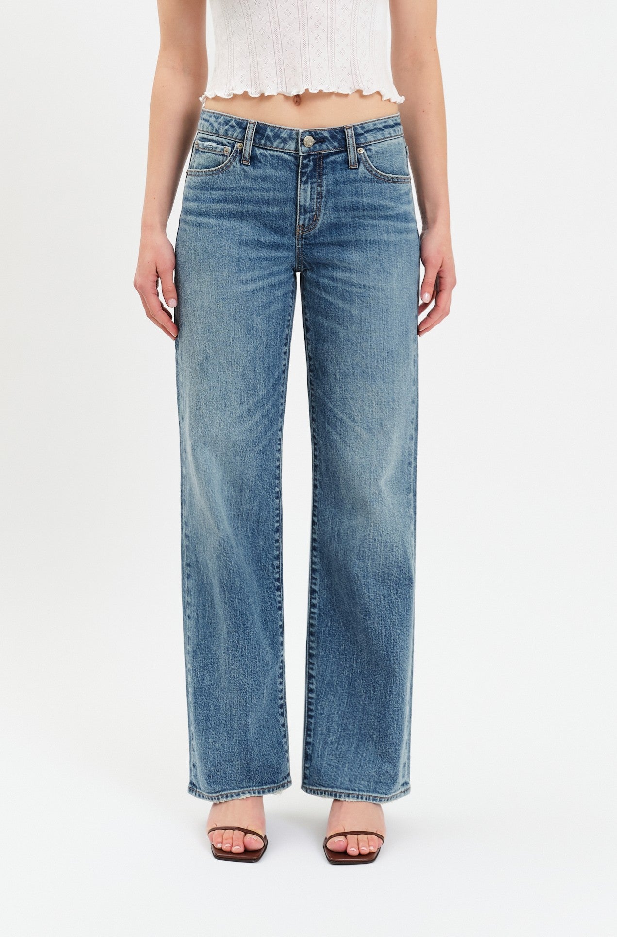 A person is shown from the torso down, wearing Daze's The Keeper Low Rise In Heartbeat Vintage jeans—a blue comfort stretch denim wide-leg style—paired with a white sleeveless crop top and black heeled sandals, standing against a plain white background.